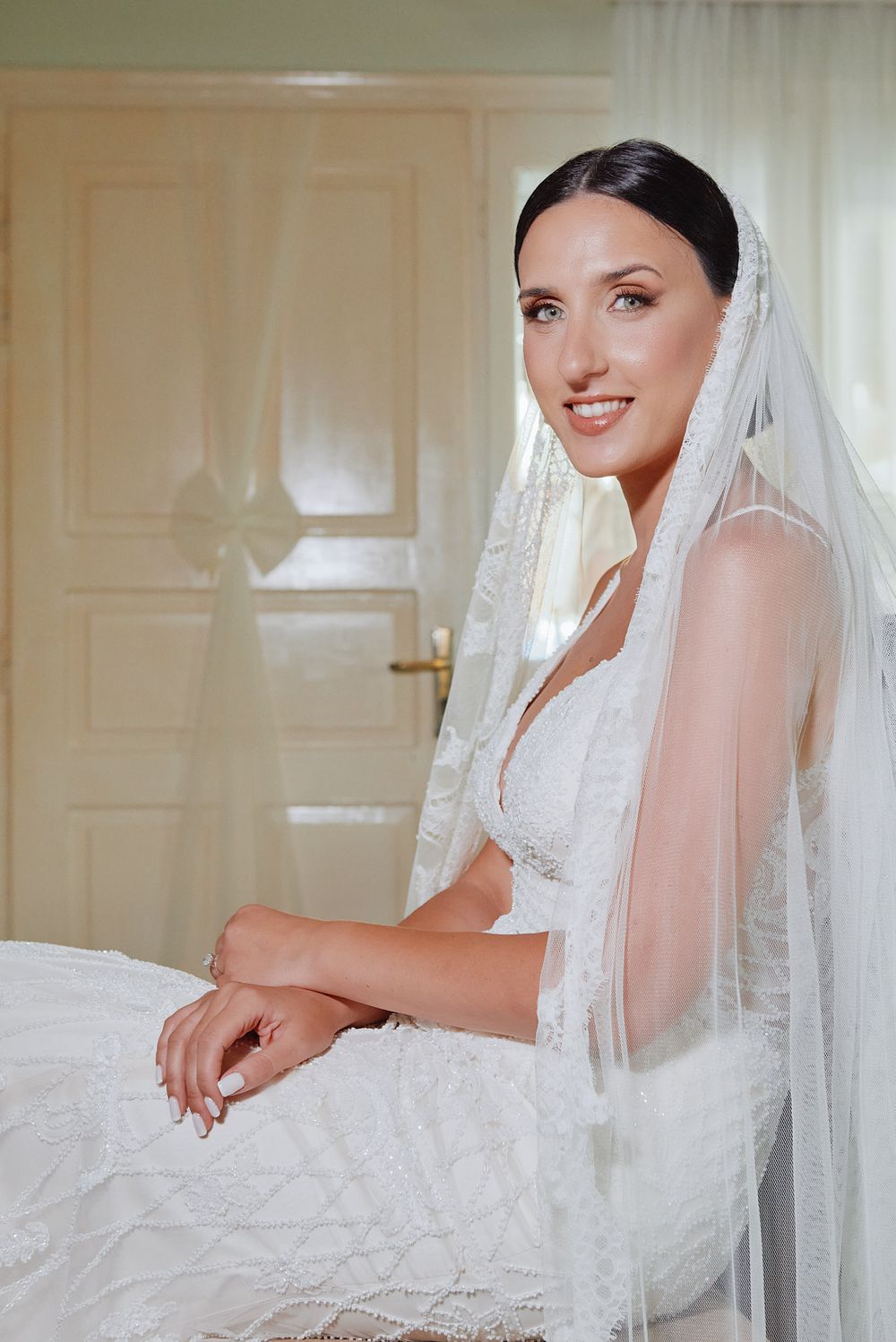 Portrait of a elegant bride in a lace wedding dress and long veil sitting indoors.
