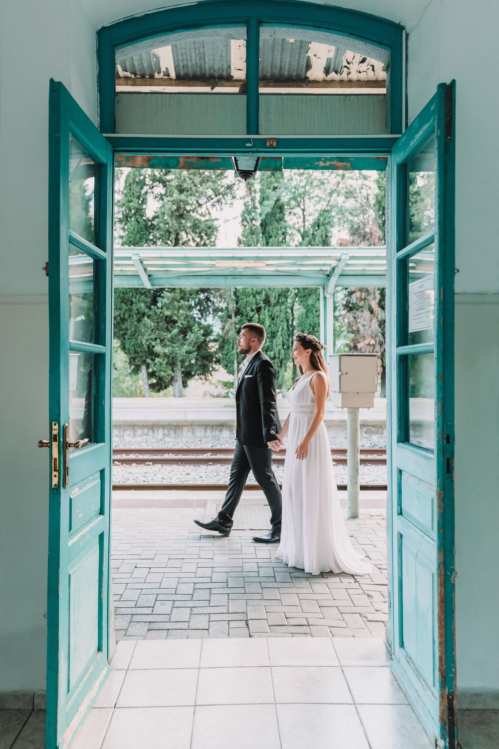 A wedding couple framed inside a doorway with vibrant teal wooden shutters in Greece.