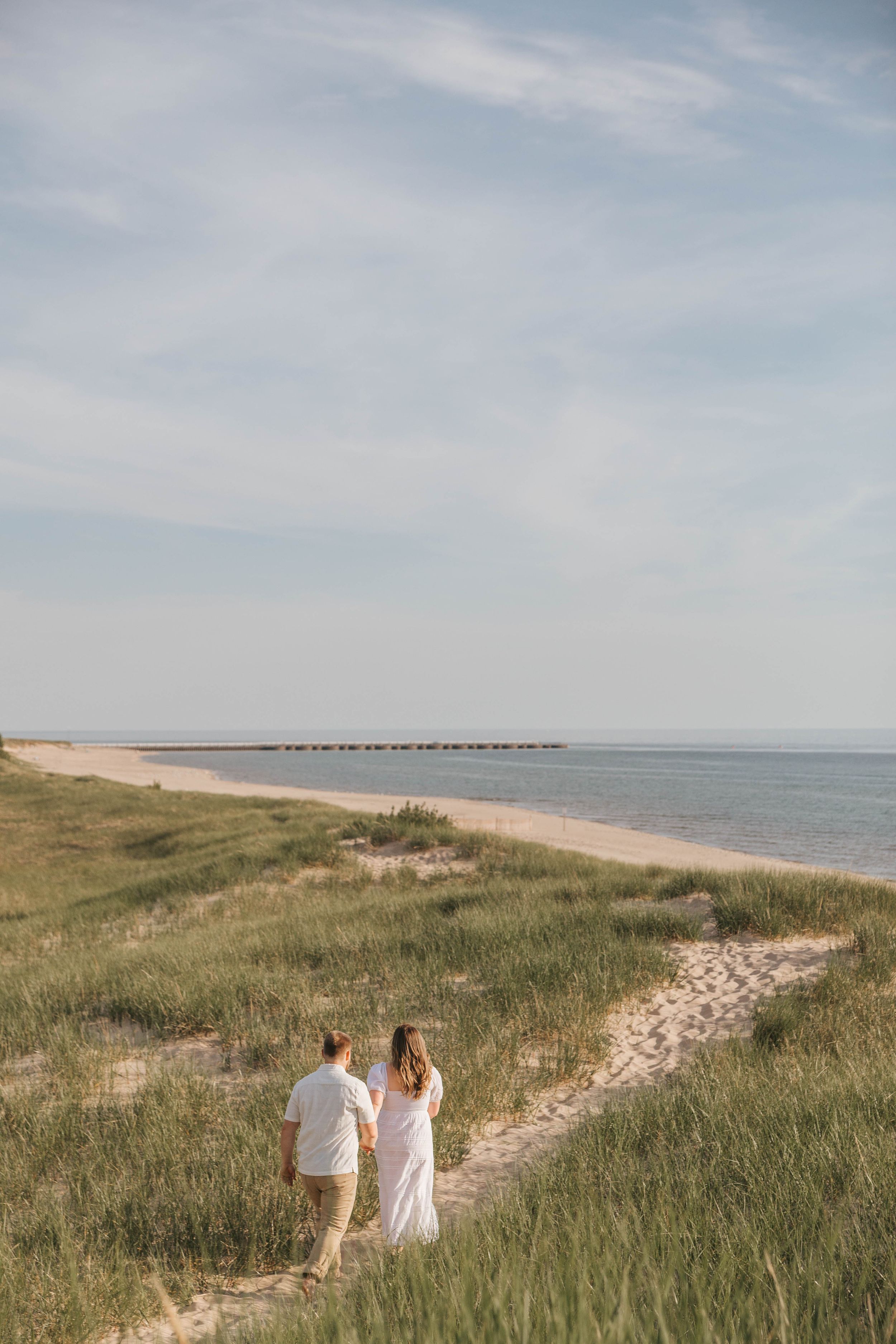 A couple in white walks hand-in-hand along a sandy beach path through grassy dunes toward the calm ocean.