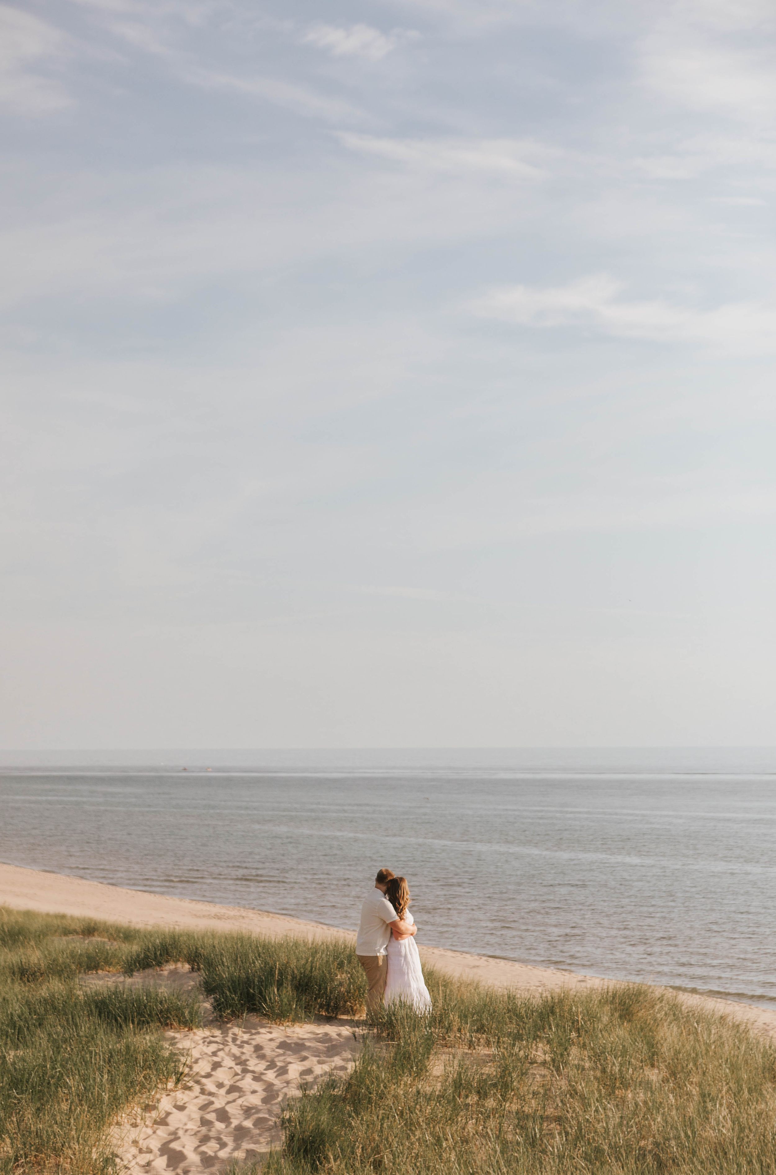 A woman in a white dress stands on sandy beach dunes overlooking a calm sea under a cloudy sky.
