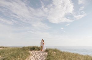 Couple embracing on sandy dune path with green grass, ocean horizon, and dramatic cloudy sky in background.