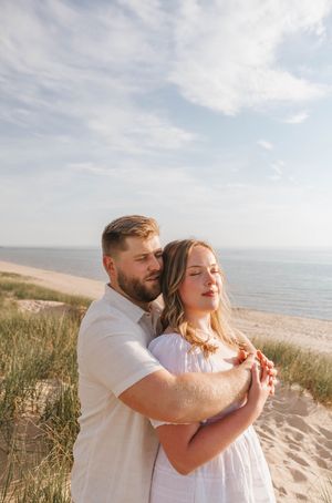 A couple embraces on a sandy beach, the man hugging the woman from behind as she closes her eyes near ocean dunes.