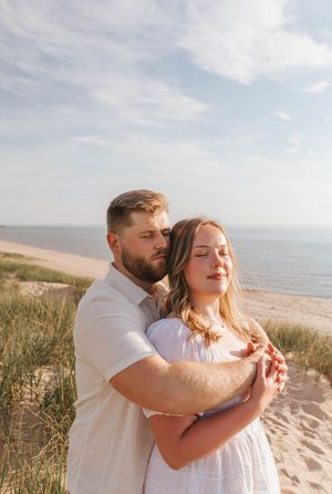 A couple embraces tenderly on a sandy beach, the man holding the woman from behind near the ocean.