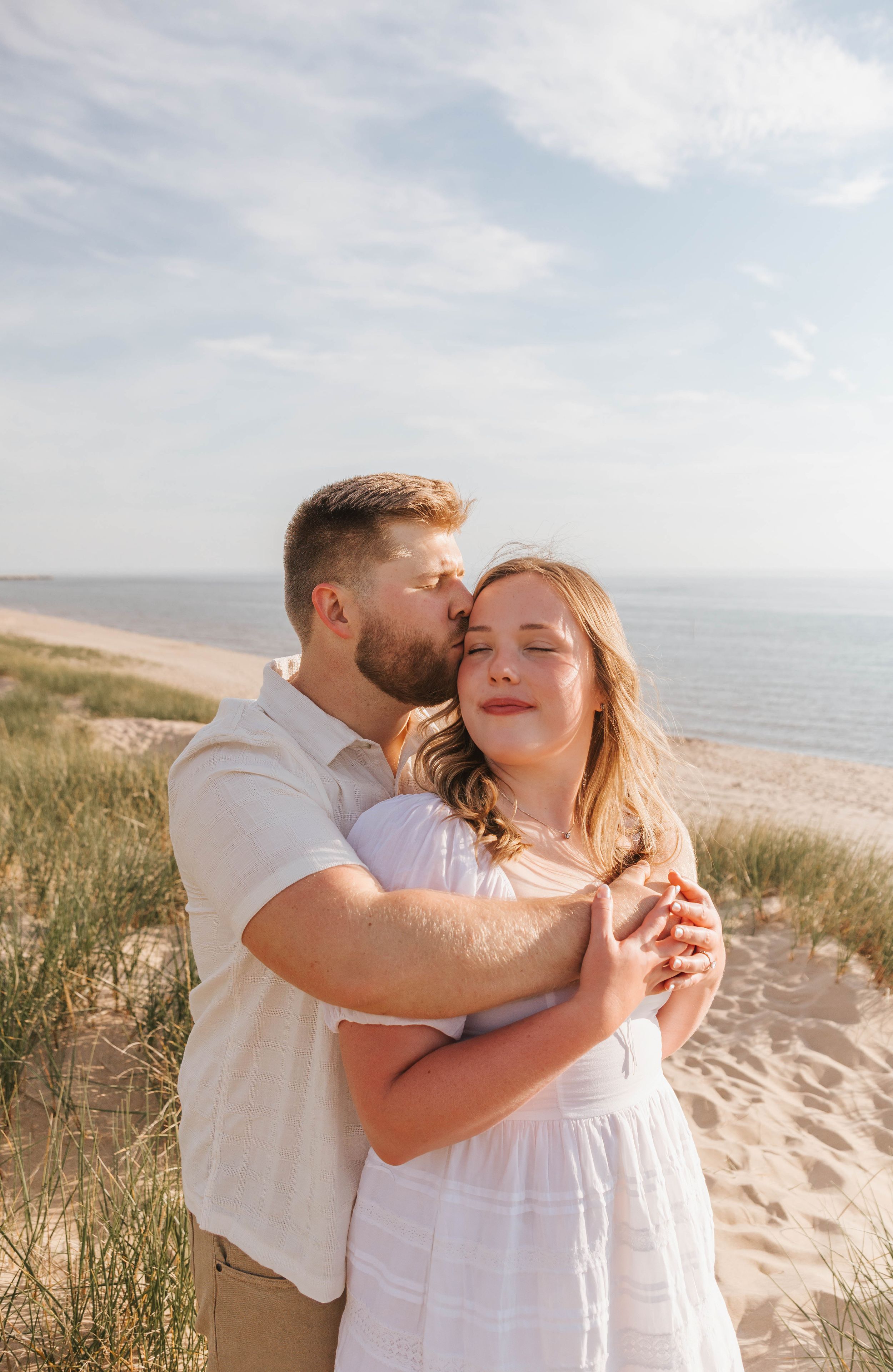 Couple embracing on sandy beach dunes, man kissing woman's cheek, both in white outfits with ocean backdrop.