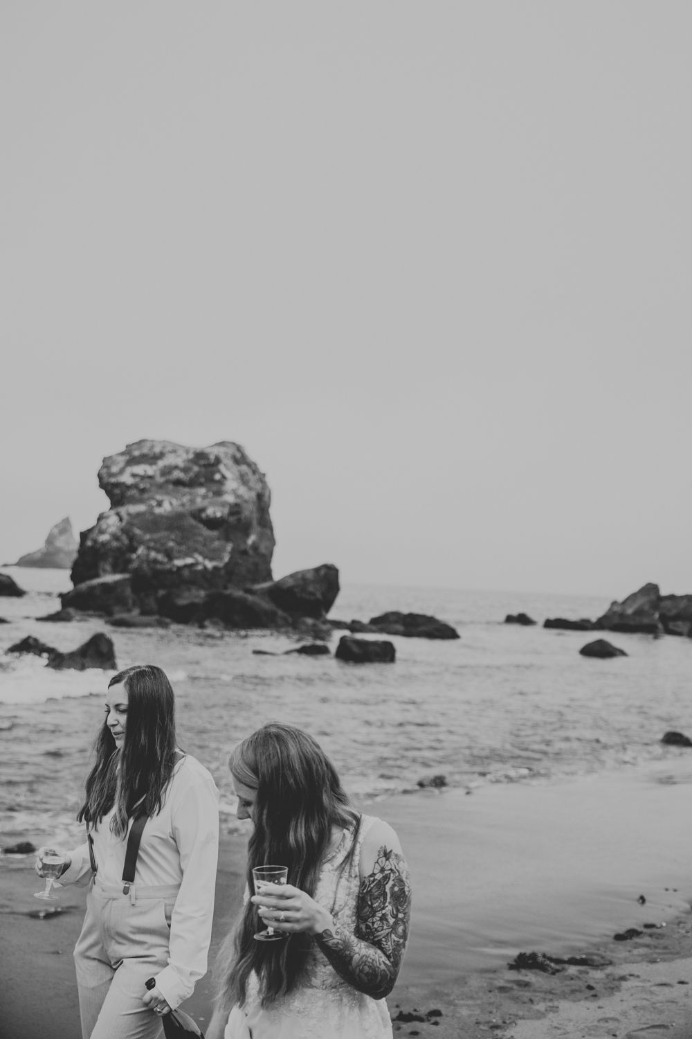 Artsy black and white photos of lesbian couple walking on the Oregon Coast holding chamgagne
