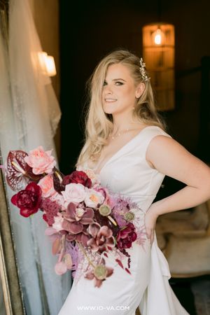Blonde bride in white gown holding a lush burgundy and pink floral bouquet, smiling softly in elegant indoor lighting.