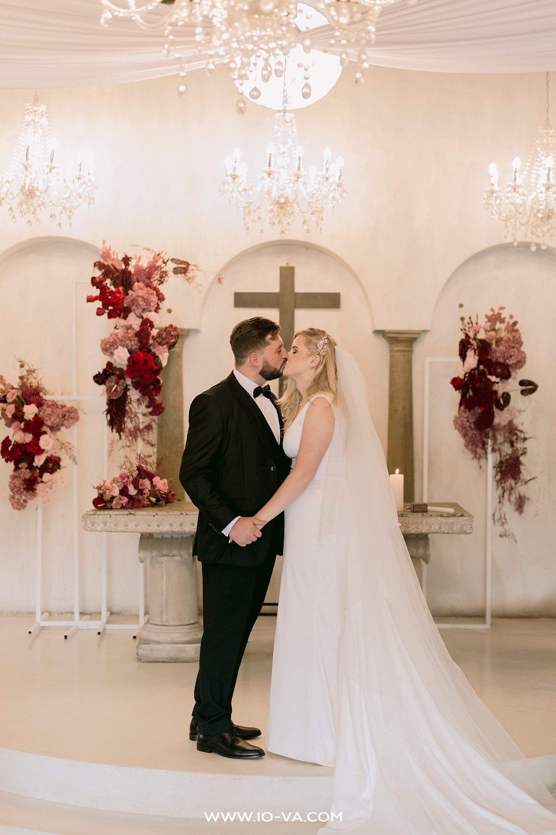Bride and groom share a kiss at a romantic chapel altar adorned with lush red and pink floral arrangements and a crystal chandelier.