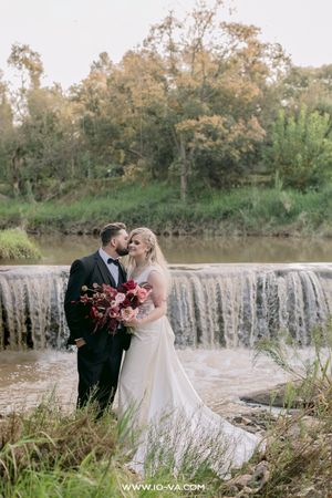 Bride and groom share a romantic kiss near a scenic waterfall, bride holding a red and pink bouquet in a white gown.