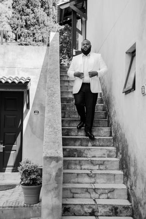 A groom in a white blazer and black pants walks down outdoor stone stairs on his wedding day.