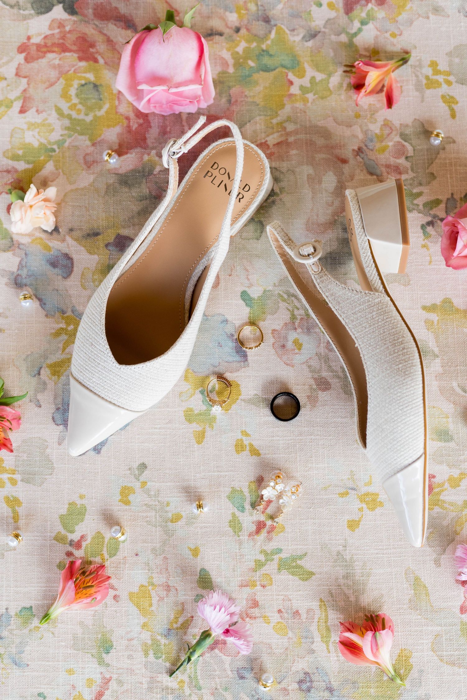 White slingback wedding heels with gold rings on floral fabric surrounded by pink roses and small flowers.