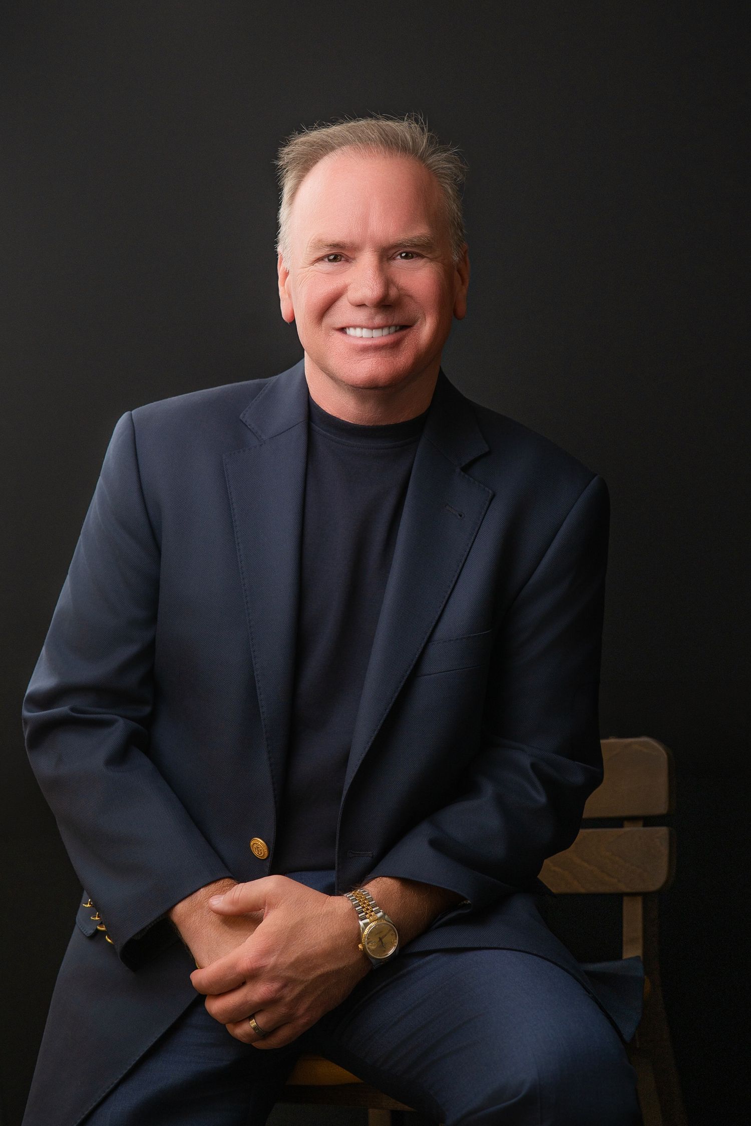 Smiling middle-aged man in navy blazer and turtleneck seated in wooden chair against dark background.