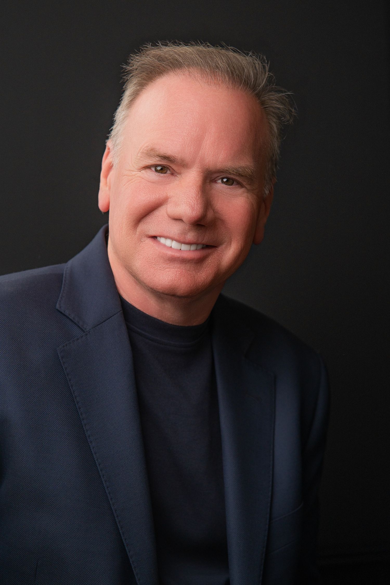Professional headshot of a smiling middle-aged man in a navy blazer and turtleneck against a dark background.