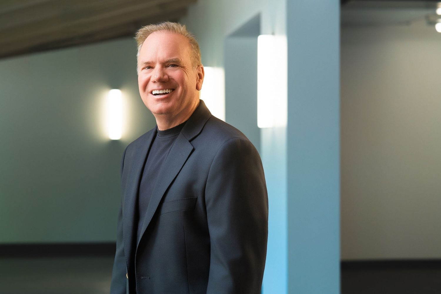 Smiling middle-aged businessman in black blazer and turtleneck standing in modern office lobby.