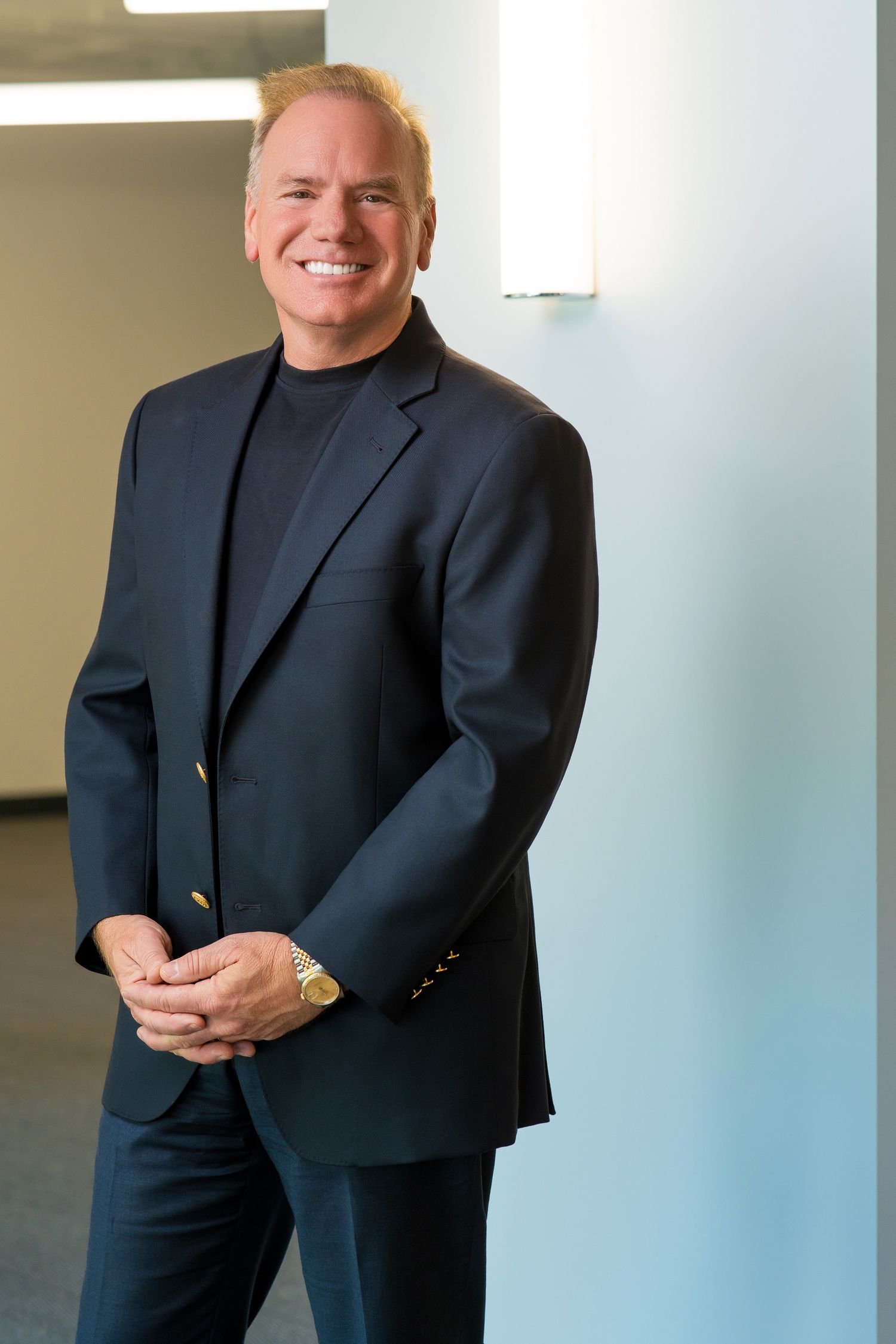Smiling middle-aged businessman in navy blazer and turtleneck stands confidently in modern office hallway.