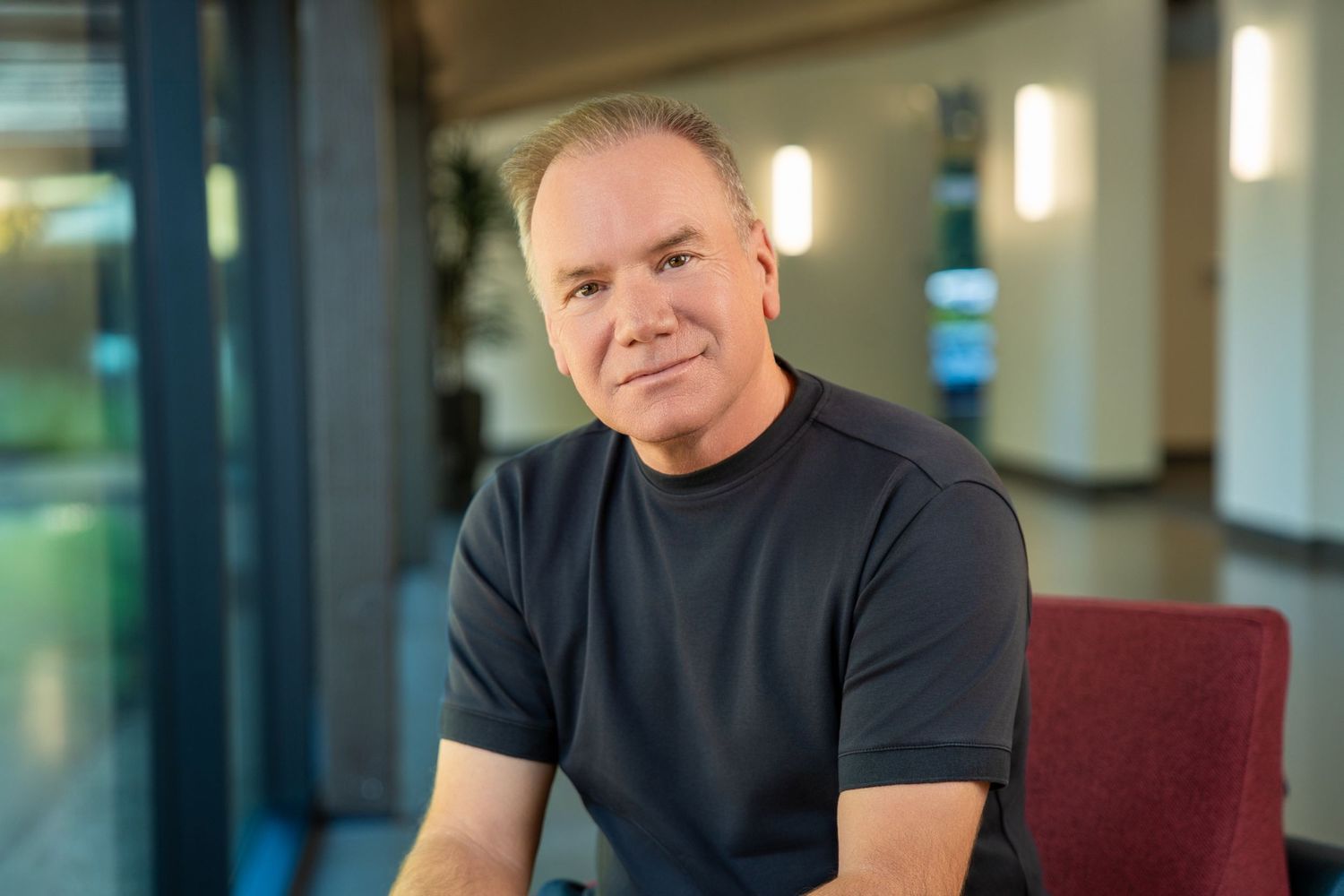 Middle-aged man in dark t-shirt smiling confidently while seated in a modern office lobby setting.