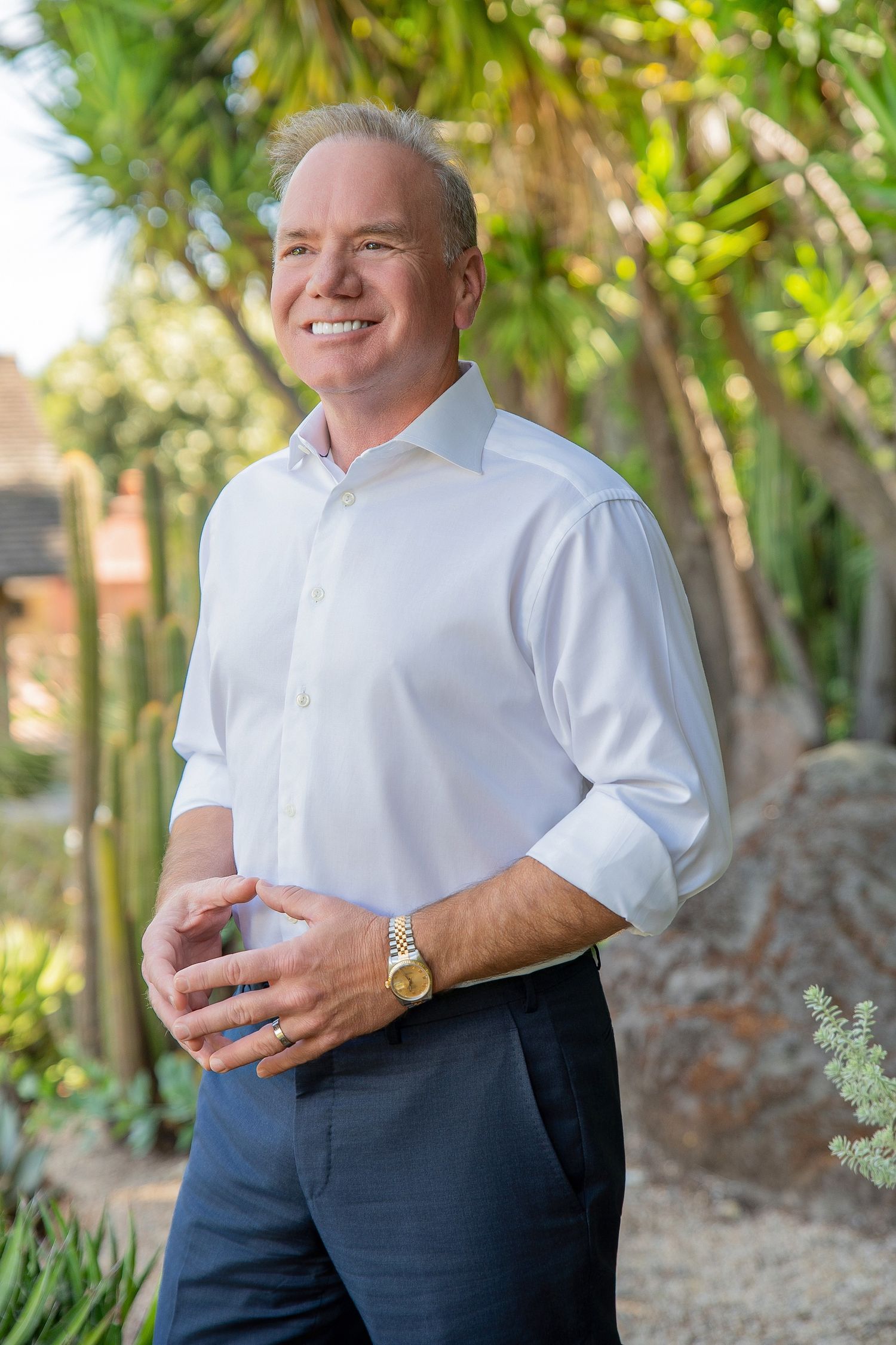Smiling man in white shirt and navy pants standing outdoors among tropical plants and palm trees.