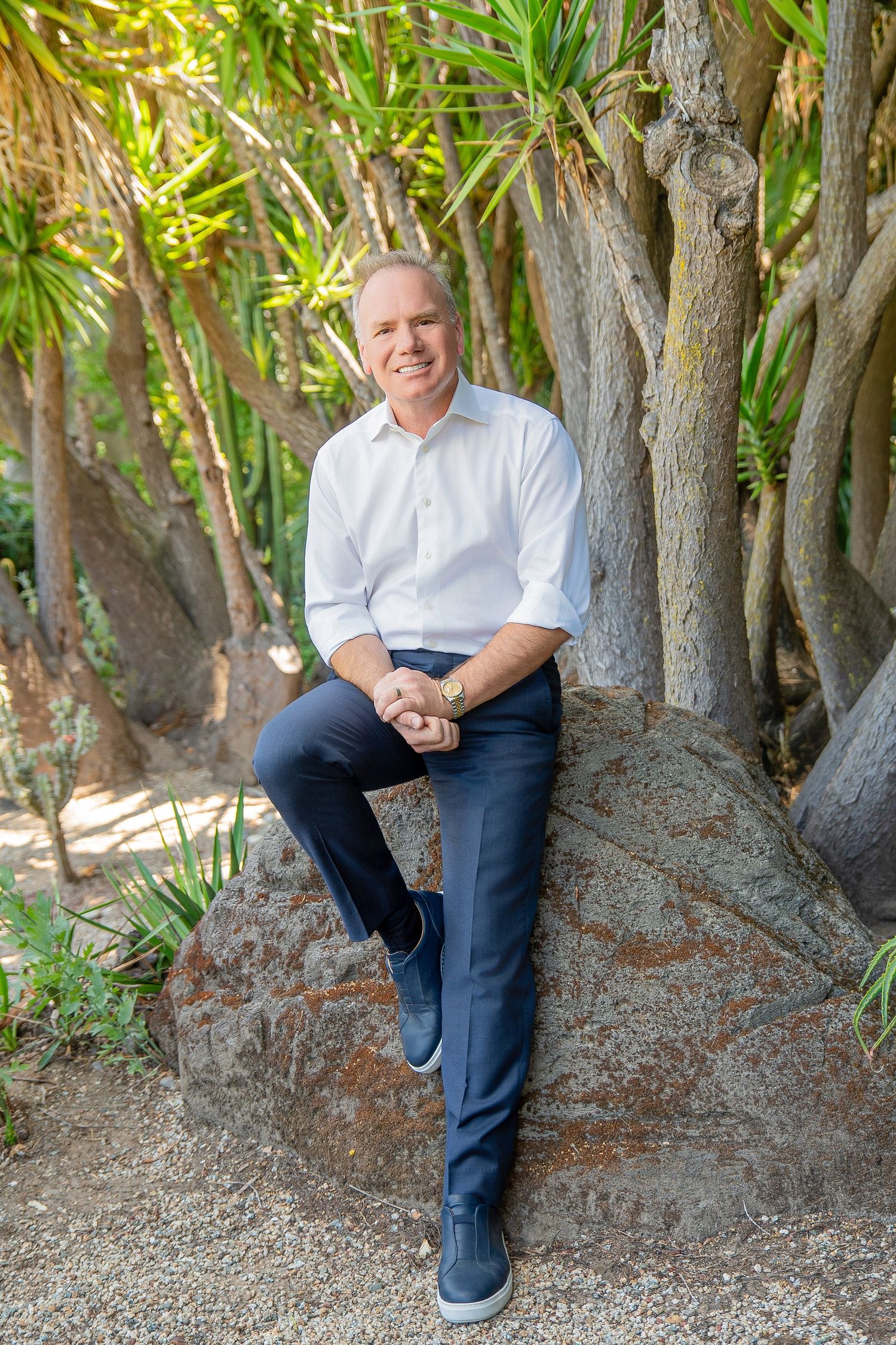 Smiling middle-aged man in white shirt and navy pants sitting on a rock surrounded by tropical trees.