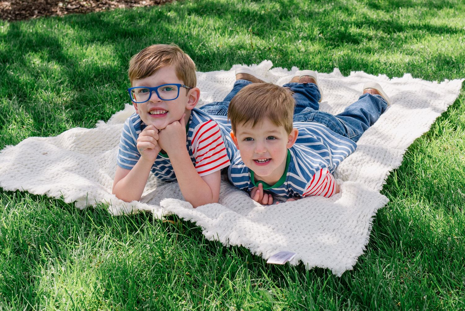 Two young boys wearing striped shirts lying on a white blanket in green grass, smiling at the camera.