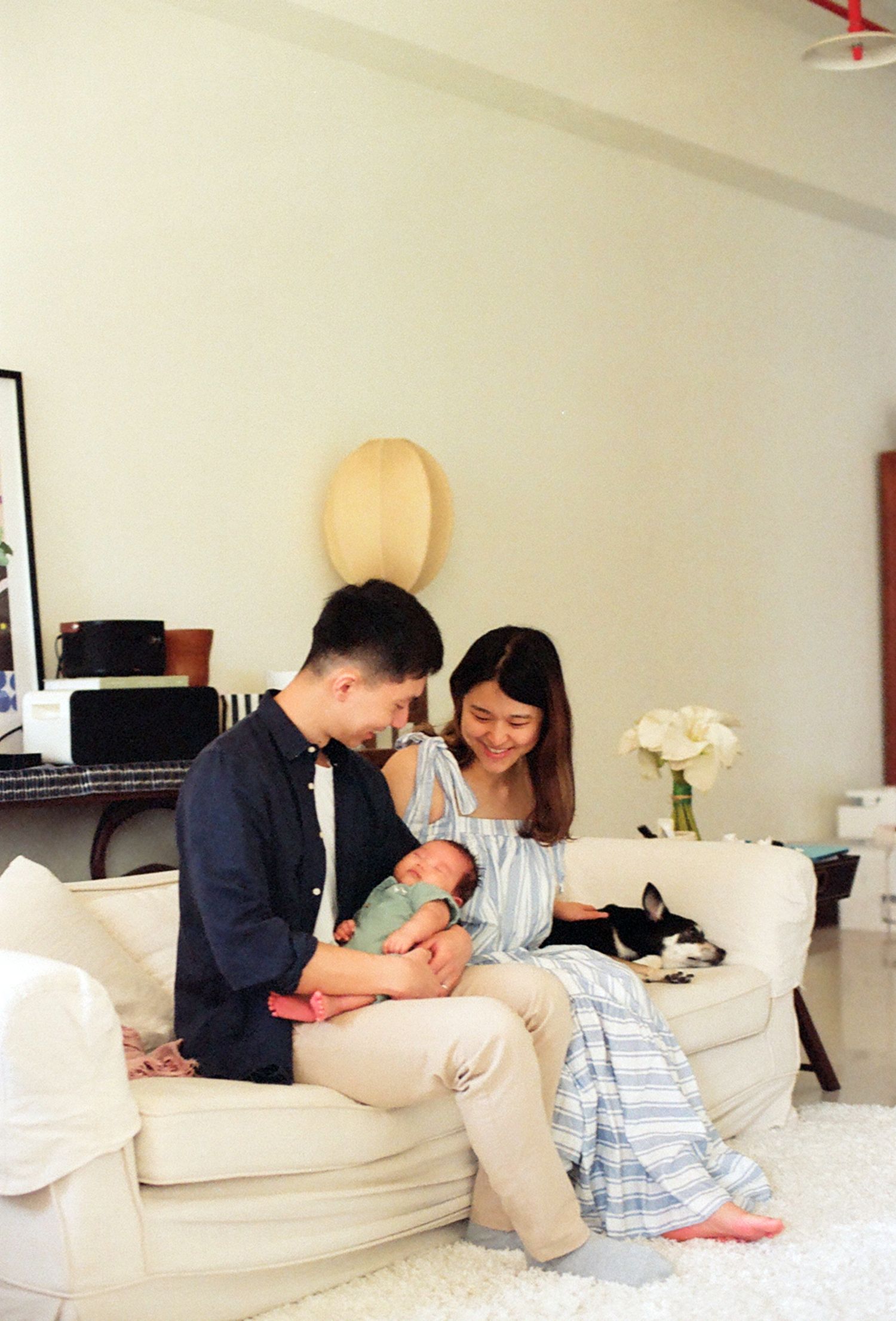 Parents smiling at their newborn baby dressed in green, with a cat resting on a bed in the background.