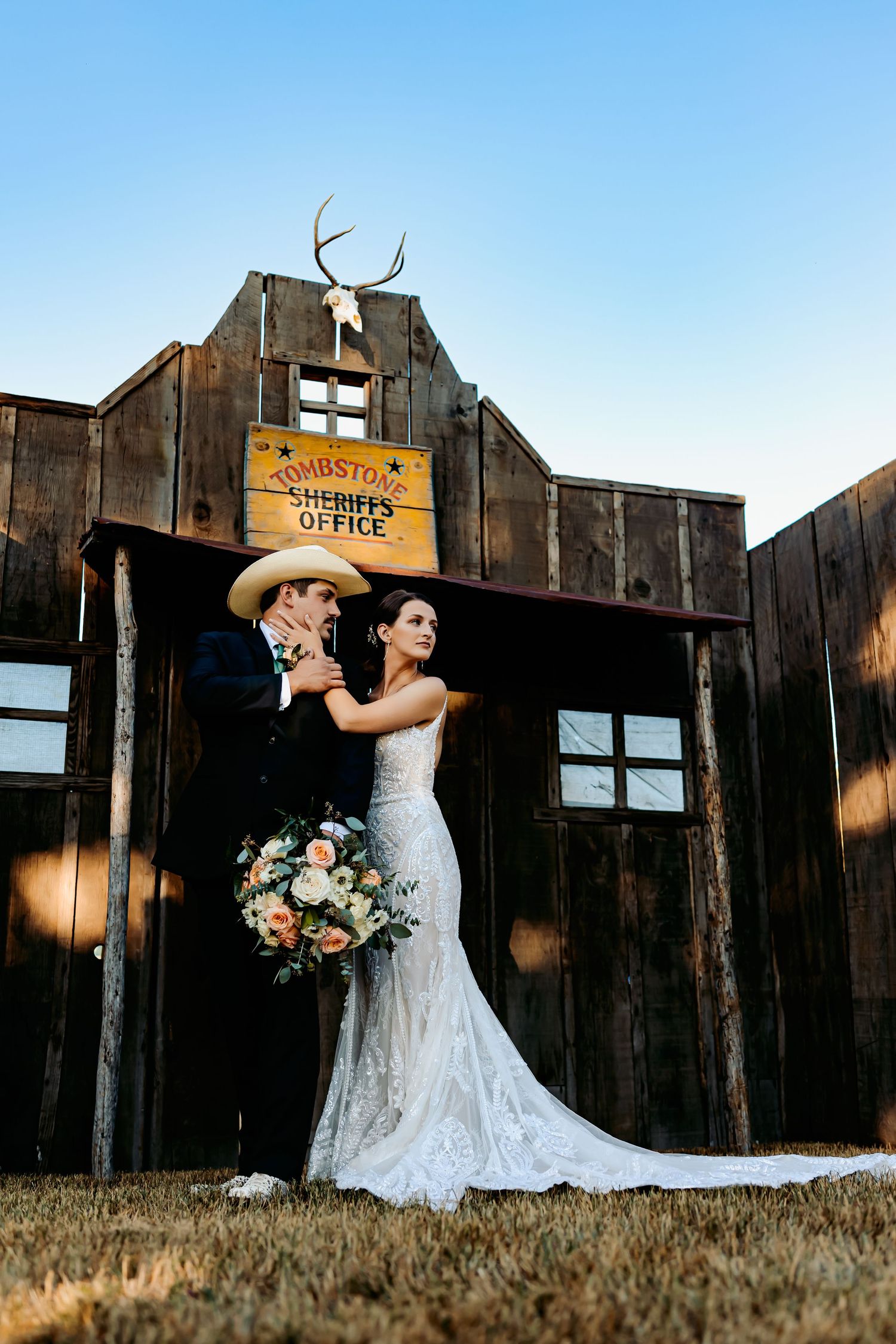 Bride and groom pose in western attire before a rustic Tombstone Sheriff's Office building at golden hour.