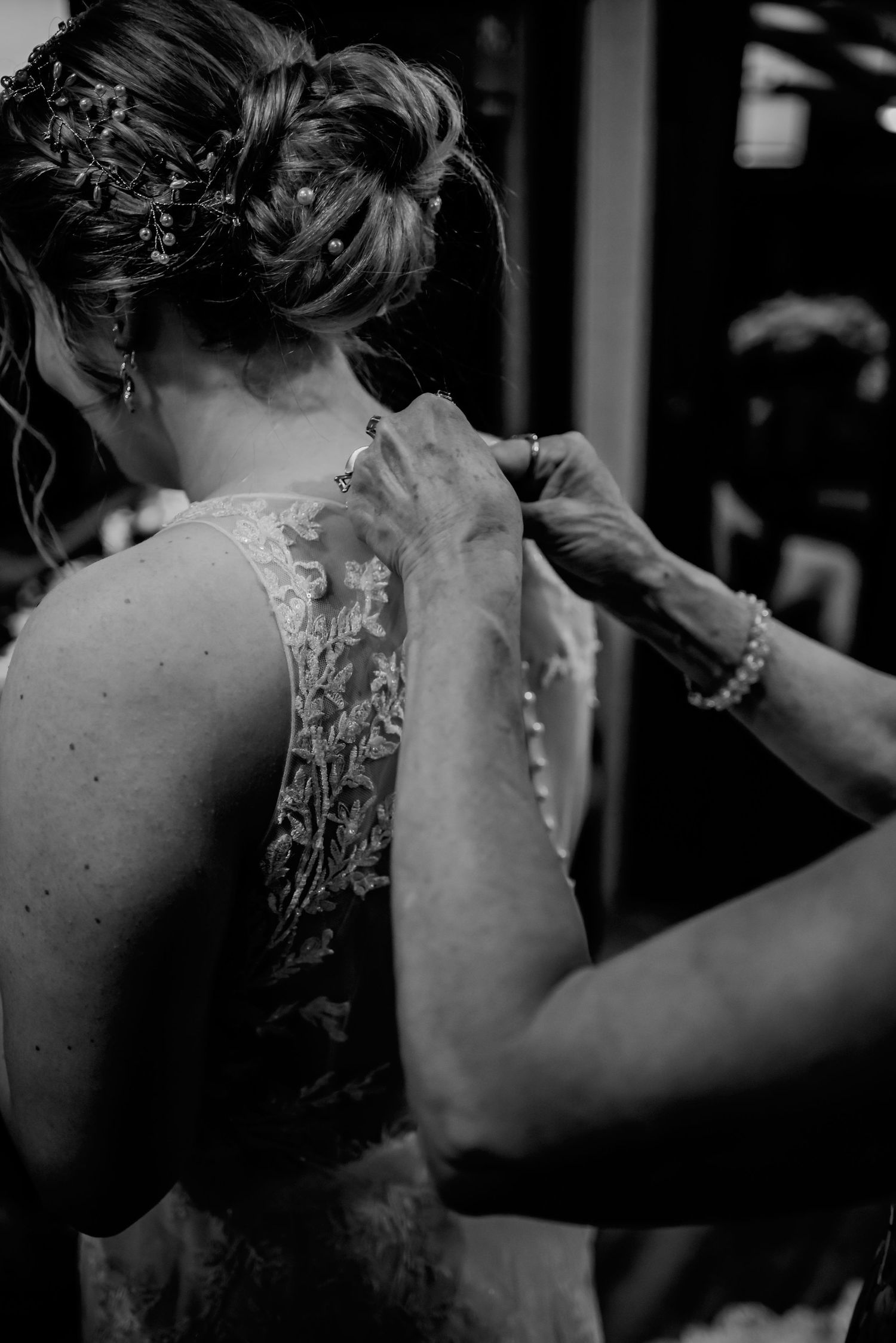 Black and white photo of a bride having her lace wedding dress fastened by a helper, with floral hair accessories.