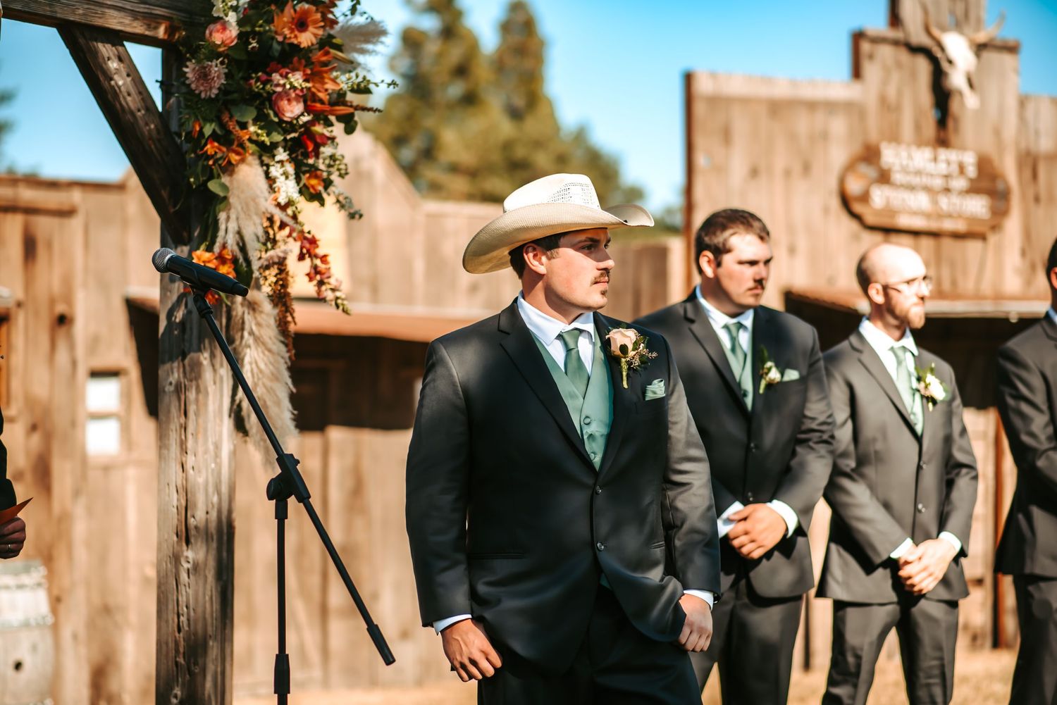 Groom in cowboy hat and dark suit stands at outdoor rustic wedding ceremony with groomsmen in background.