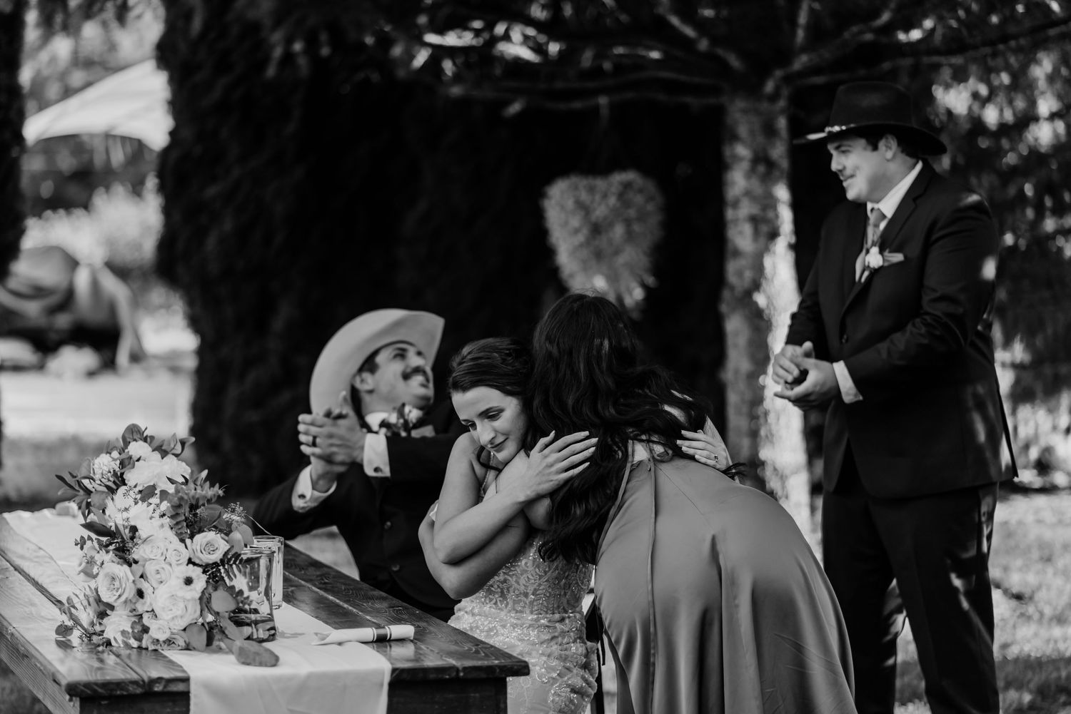 Black and white wedding photo of bride receiving an emotional hug while guests celebrate at an outdoor rustic ceremony.