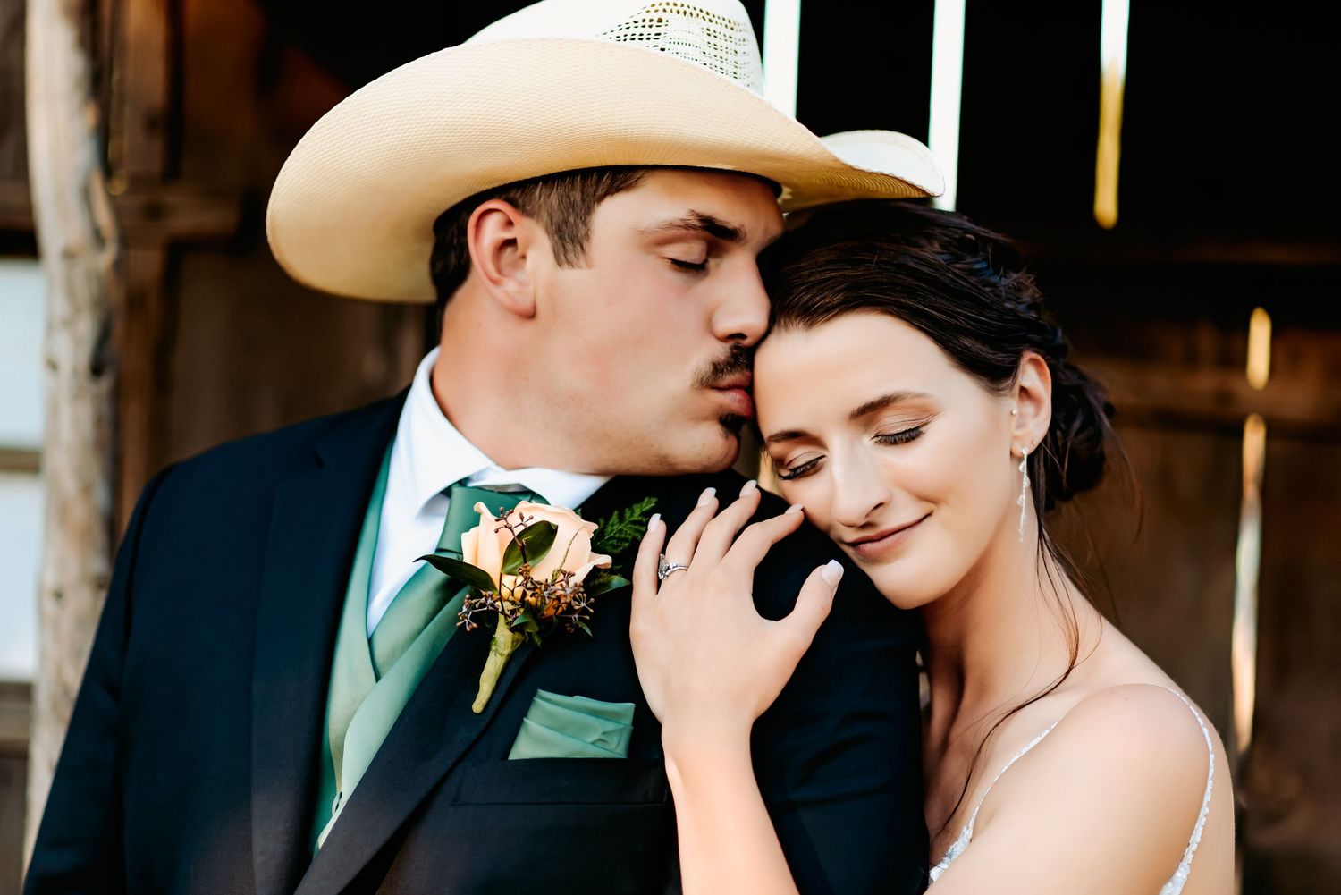 Groom in cowboy hat and green tie kisses bride's temple during romantic rustic wedding portrait.