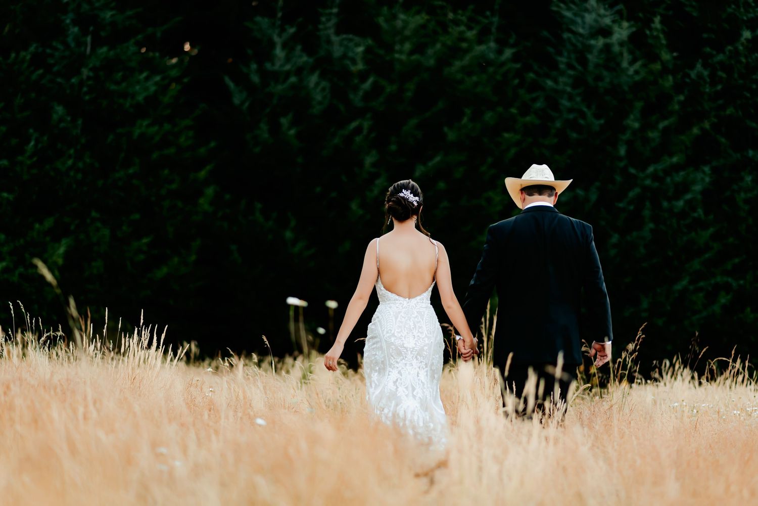 Bride in white lace gown and groom in cowboy hat walk hand-in-hand through golden wheat field.
