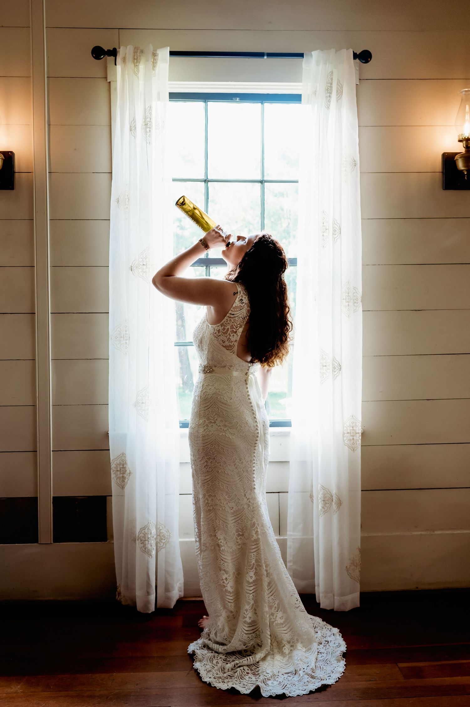 Bride in lace wedding gown drinking champagne from bottle by sunlit window on wedding day.