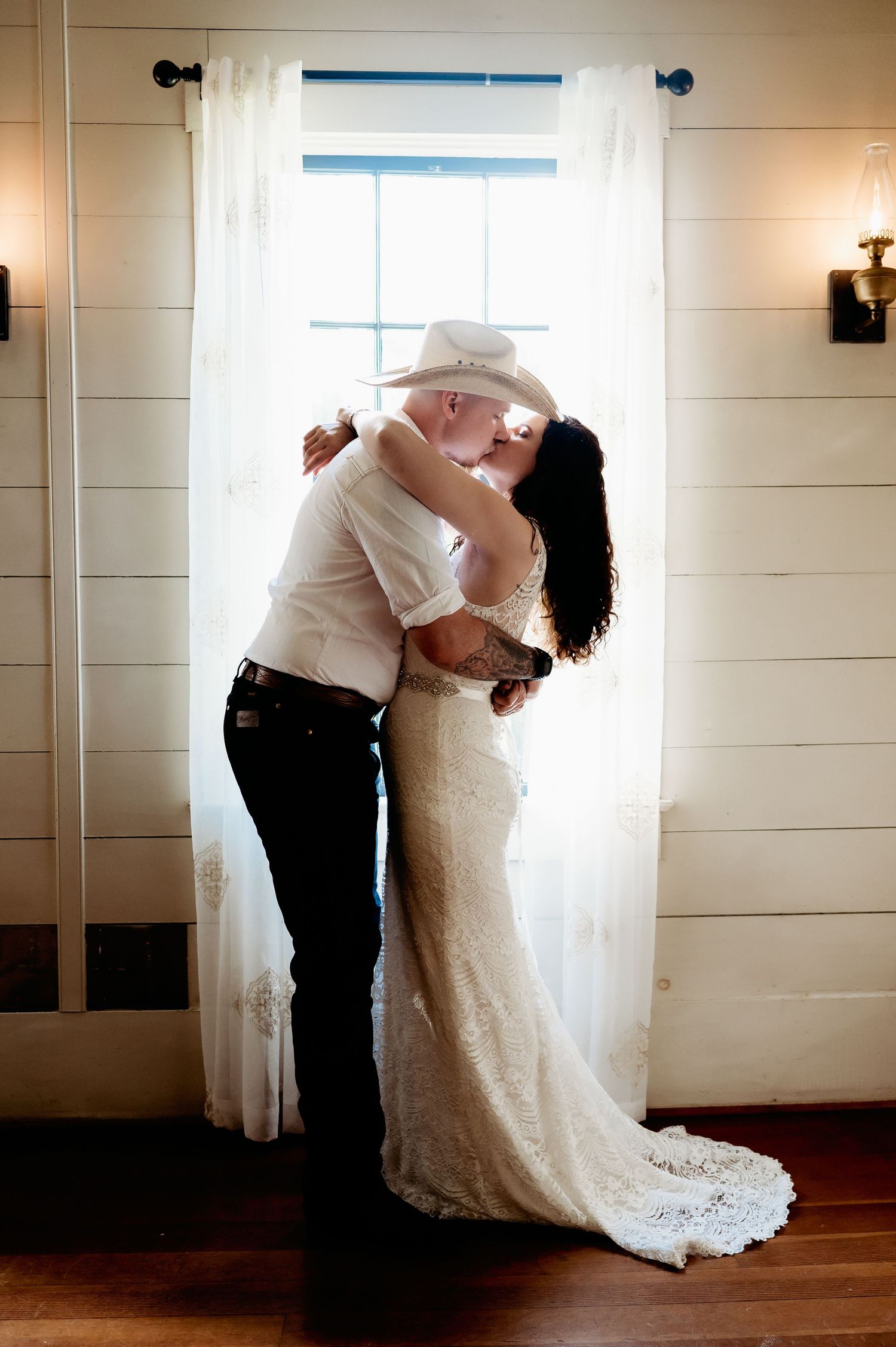 Bride and groom sharing a romantic kiss in front of a sunlit window, bride in lace gown, groom in white shirt and cowboy hat.