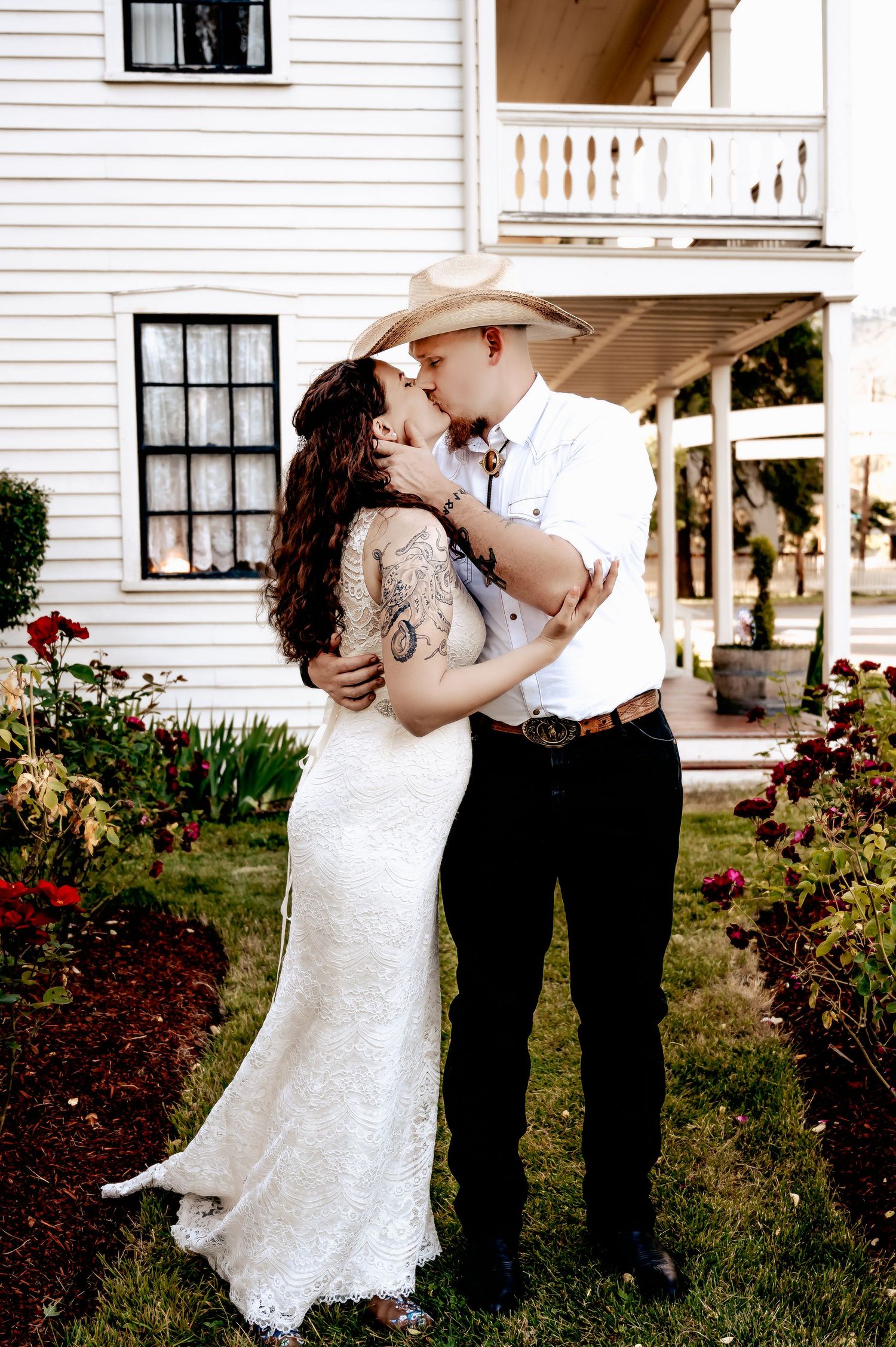 A couple shares a romantic kiss in a garden, bride in lace gown and groom in cowboy hat, beside a white farmhouse.