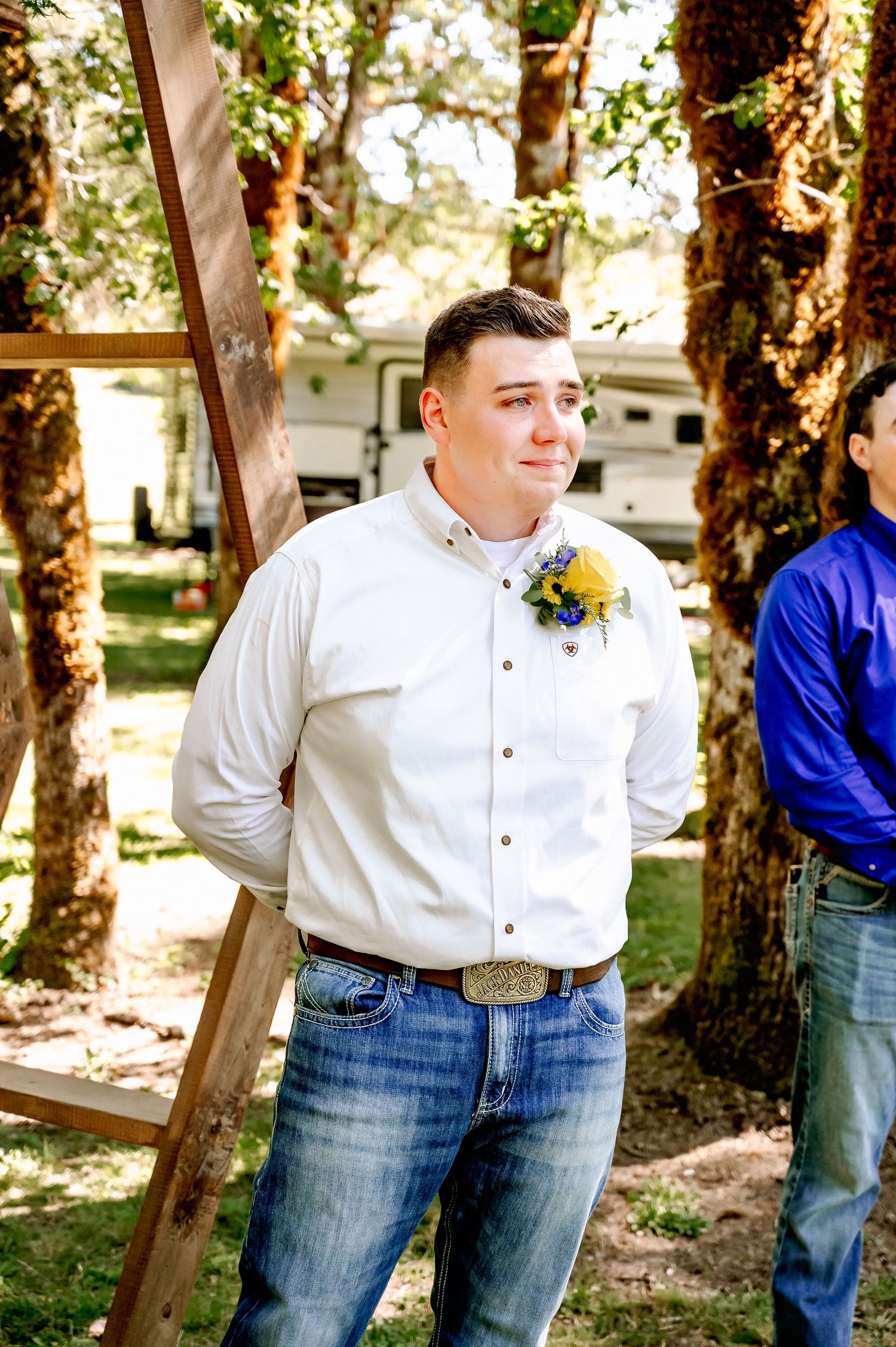 Groom in white shirt with floral boutonniere and jeans stands outdoors at rustic wedding ceremony.