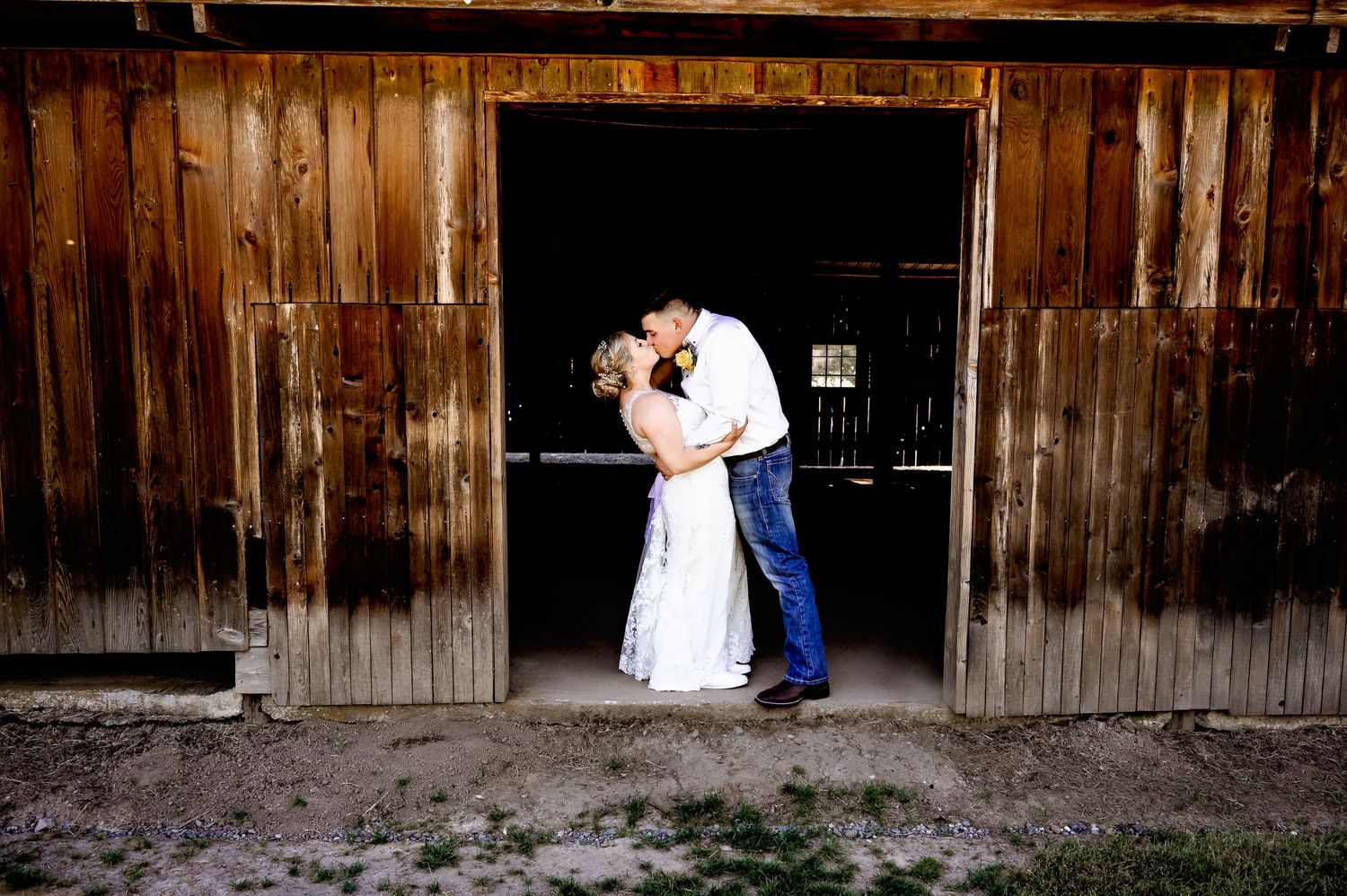 Couple sharing a romantic kiss in a rustic barn doorway, bride in white gown and groom in blue jeans.