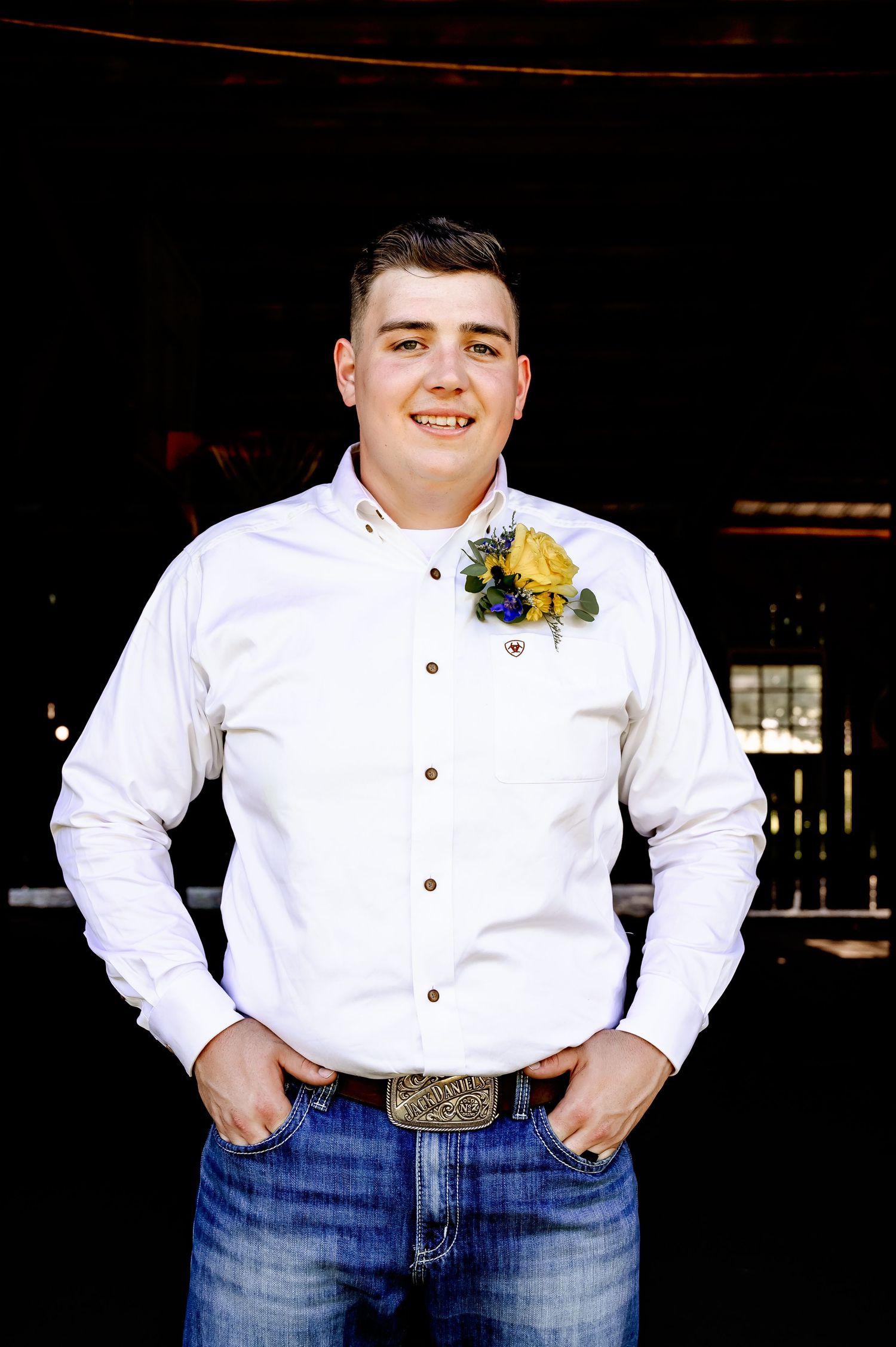 Young groom in white shirt with yellow boutonniere and jeans poses in rustic barn setting.