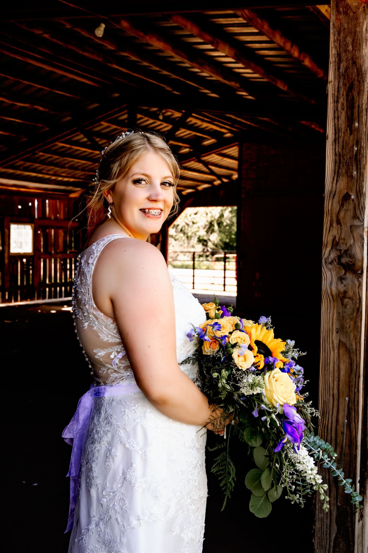 Bride in lace gown holds colorful yellow and purple bouquet, smiling in rustic barn doorway.