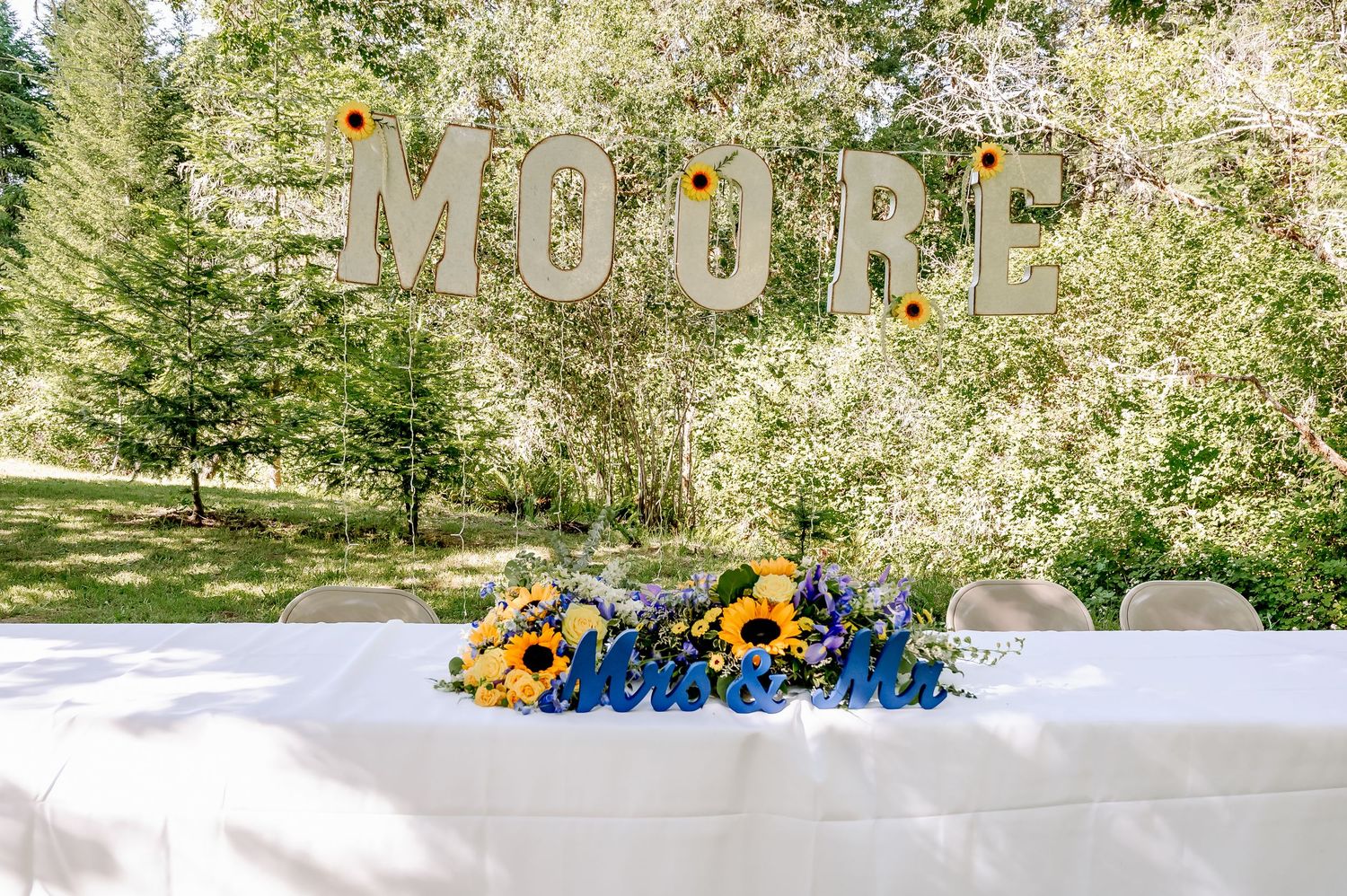 Outdoor wedding reception table with sunflowers beneath large MOORE marquee letters surrounded by lush green trees.