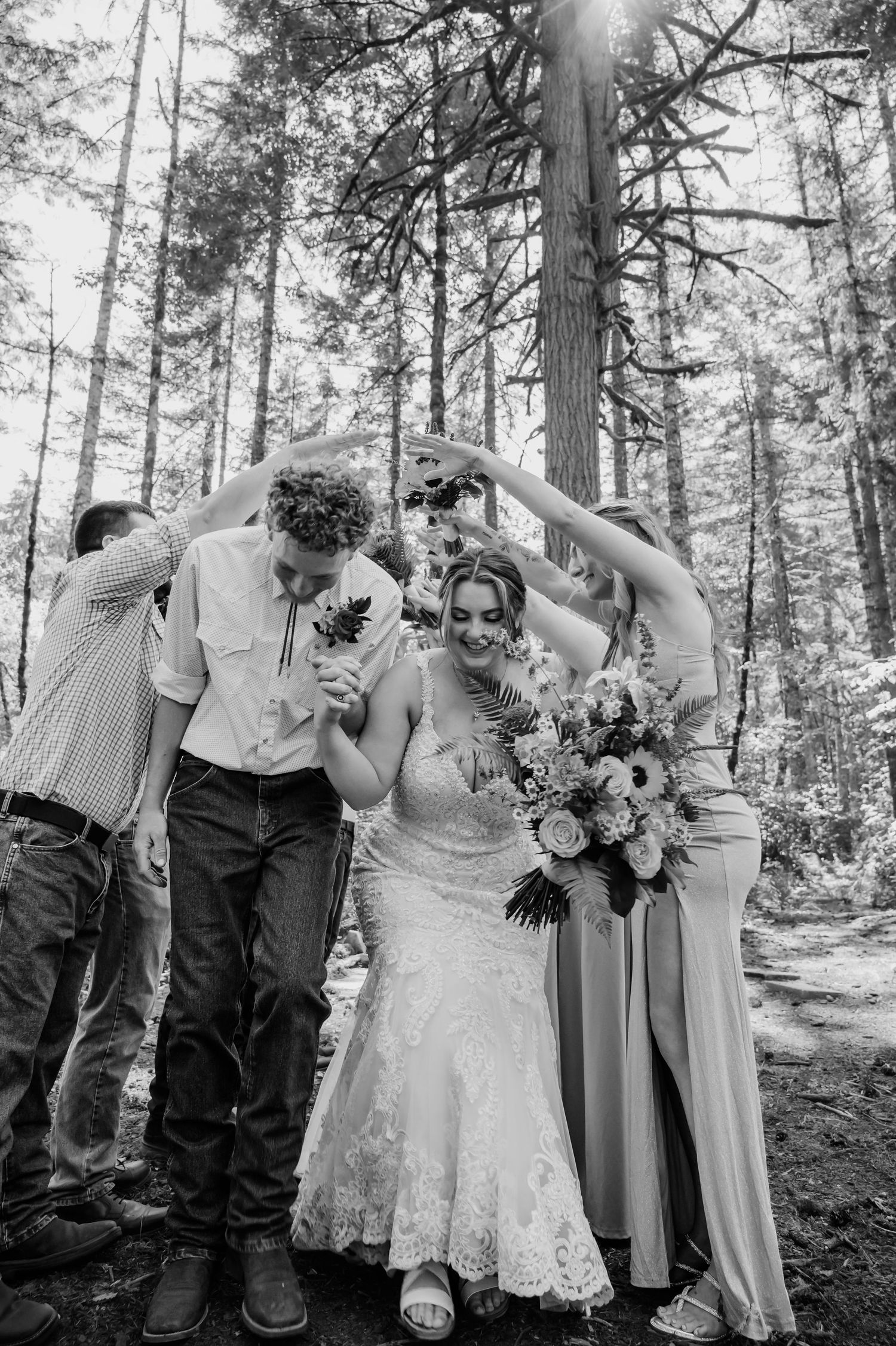 Black and white photo of a bride and groom celebrating with wedding party in a forest setting.