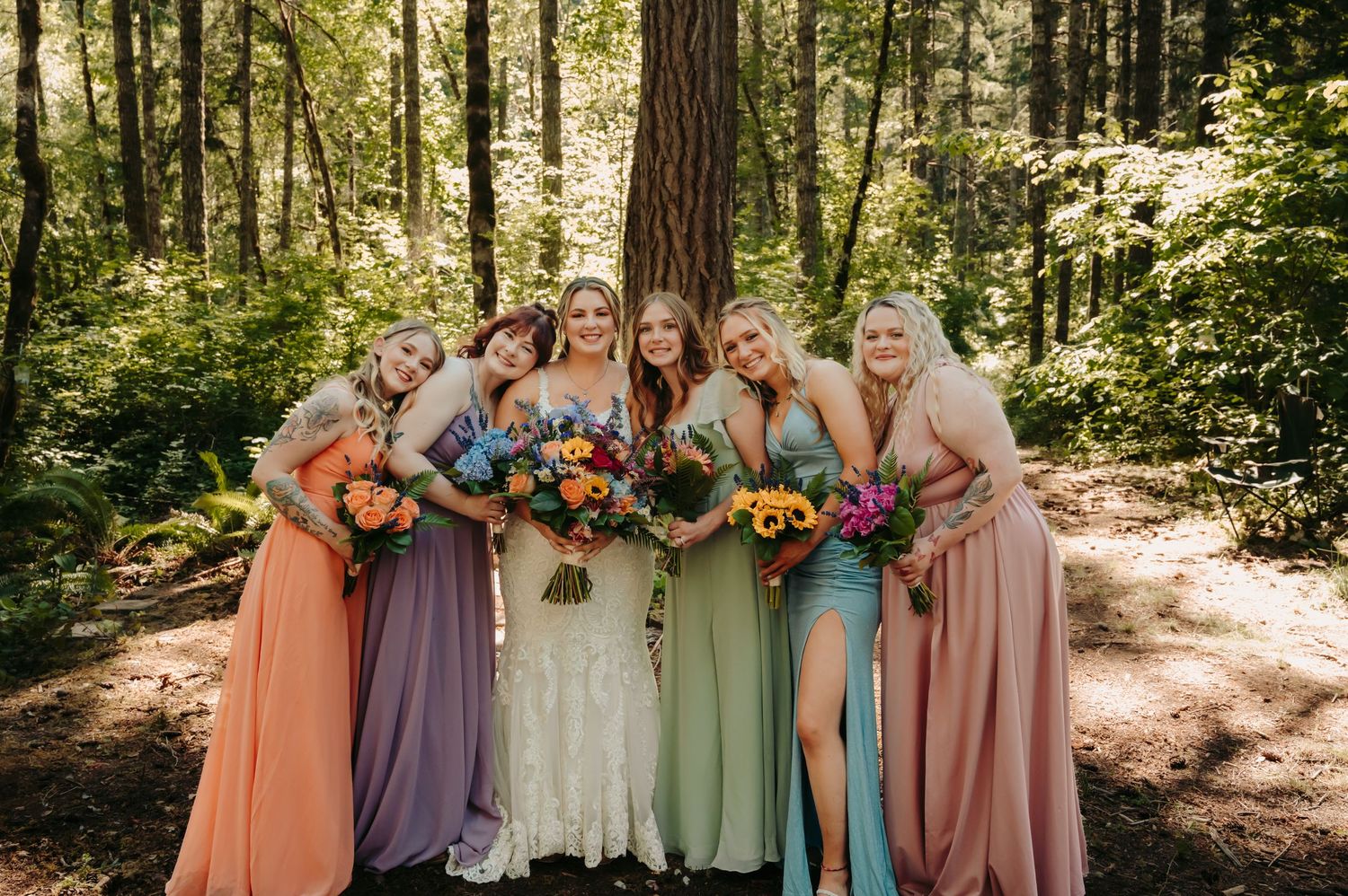 Bride and bridesmaids in colorful mismatched gowns holding bouquets, posing together in a lush forest setting.