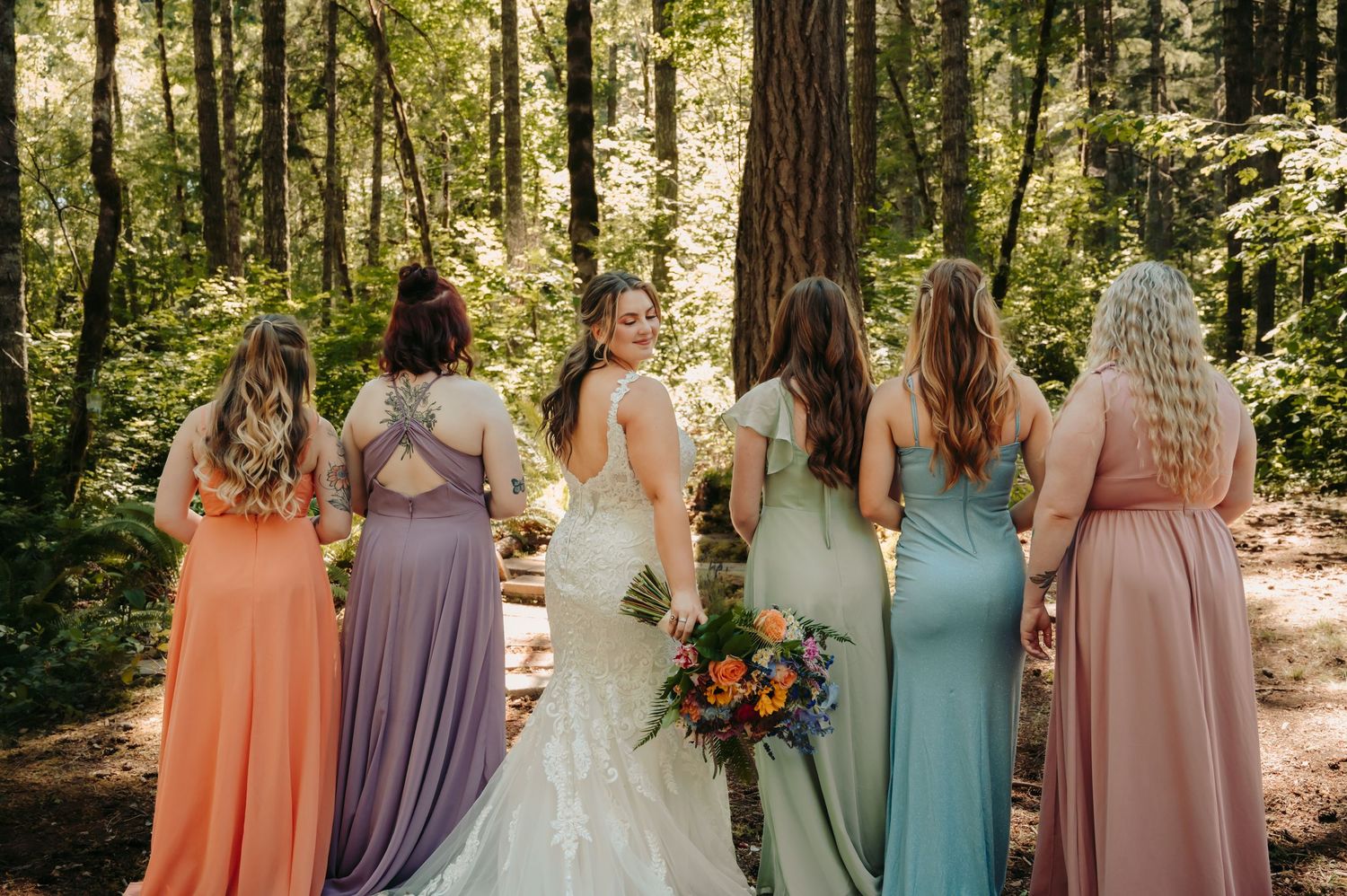 Bride in white lace gown stands with five bridesmaids in colorful mismatched dresses in a sunlit forest setting.