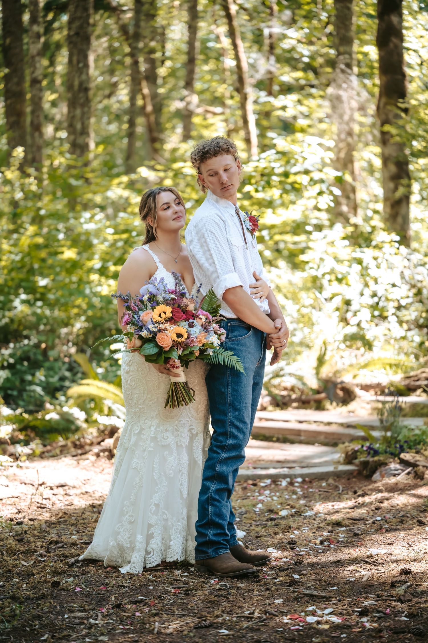 Bride in lace gown holding colorful bouquet stands with groom in white shirt and jeans in a sunlit forest setting.