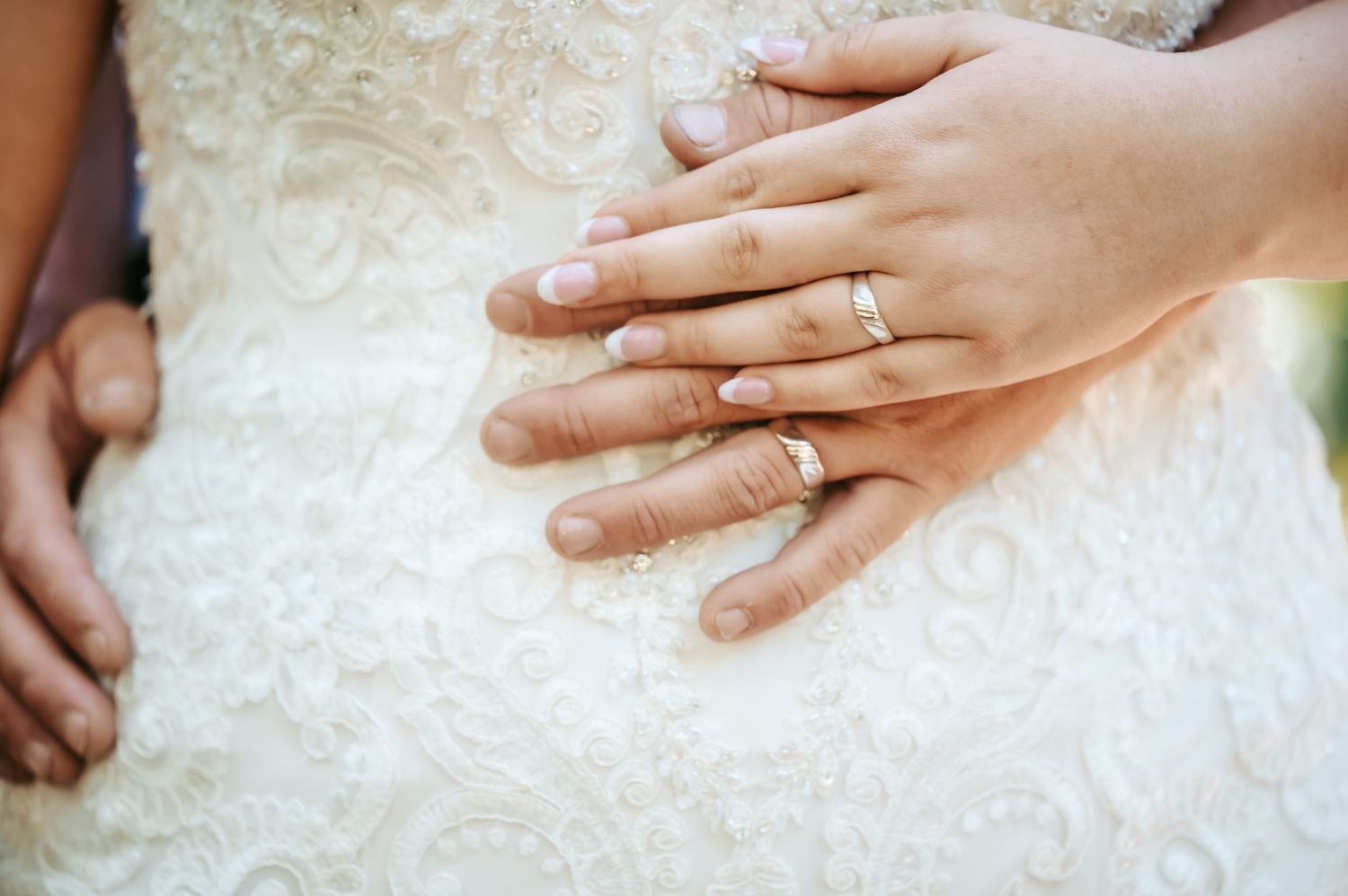 Close-up of newlywed couple's hands wearing gold wedding bands resting on white lace wedding dress.