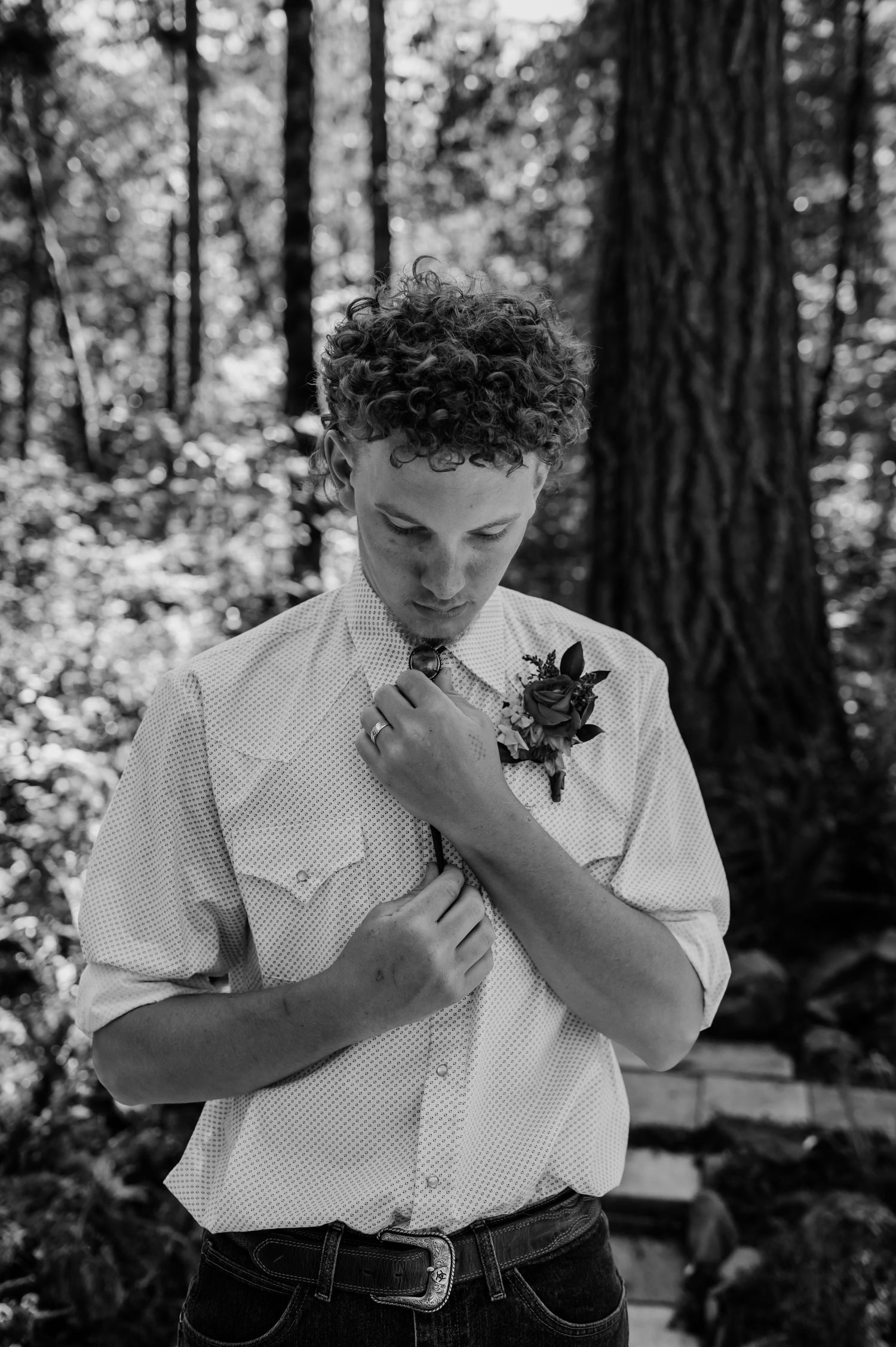 Groom in white shirt adjusting boutonniere in forest setting, black and white wedding photo.