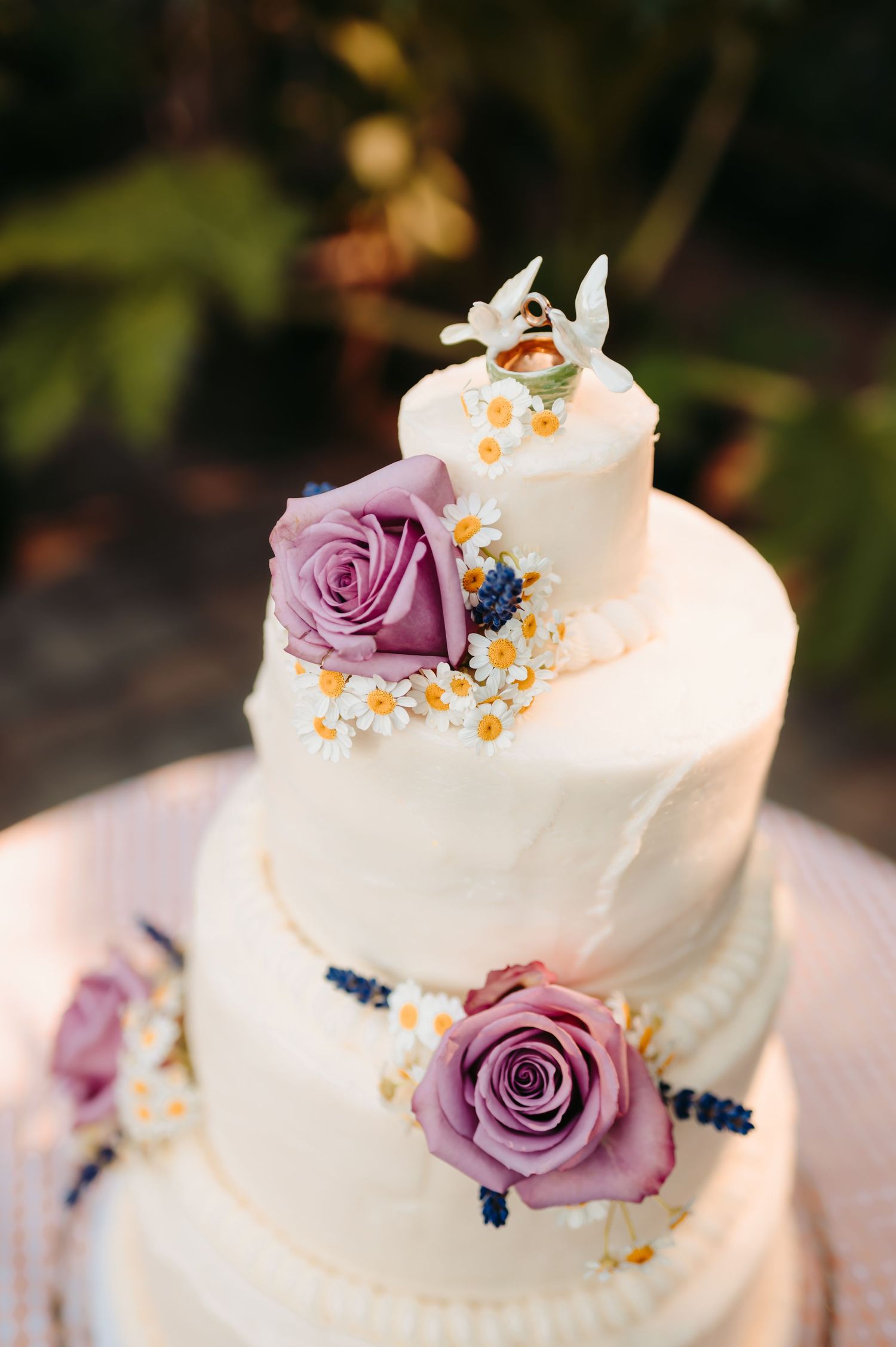 Three-tier white wedding cake adorned with purple roses and delicate daisy flowers.