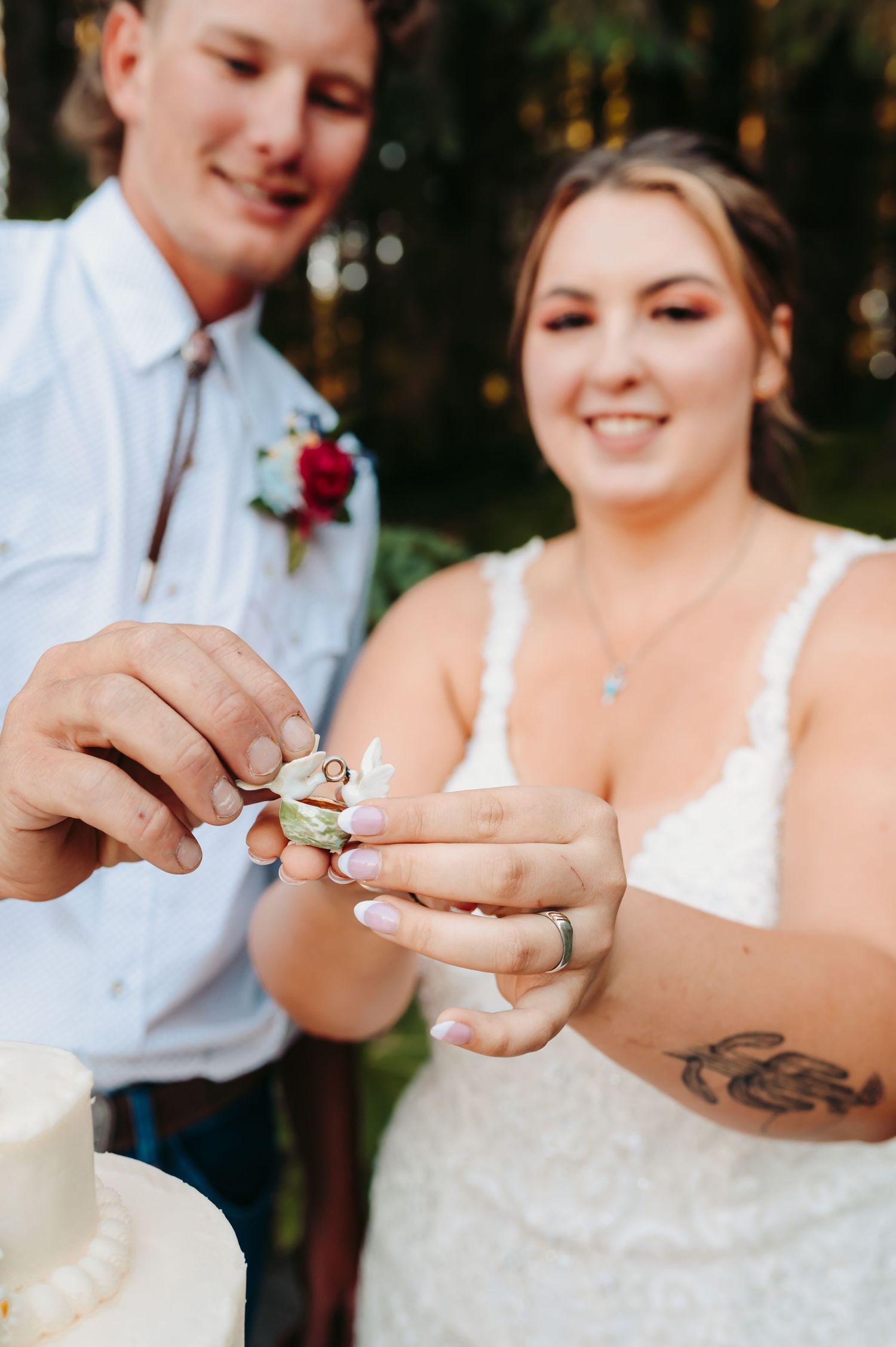 Bride and groom sharing wedding cake, smiling couple in lace dress and white shirt with boutonniere at outdoor reception.