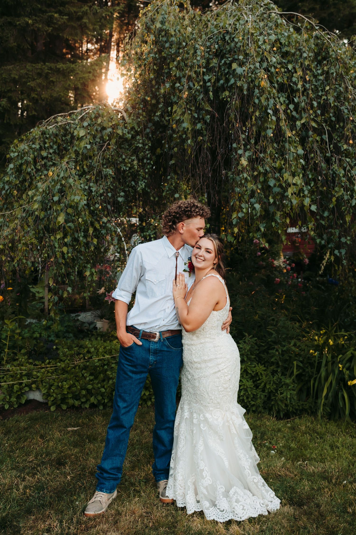 Bride and groom embracing under weeping willow tree at golden hour outdoor wedding.