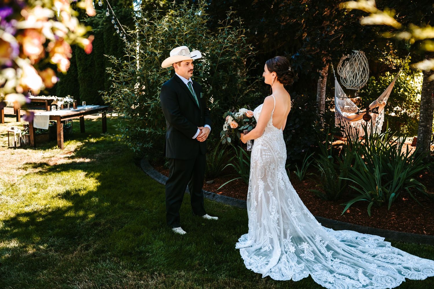 Bride in lace wedding gown with train shares first look moment with man in black suit outdoors in garden setting.