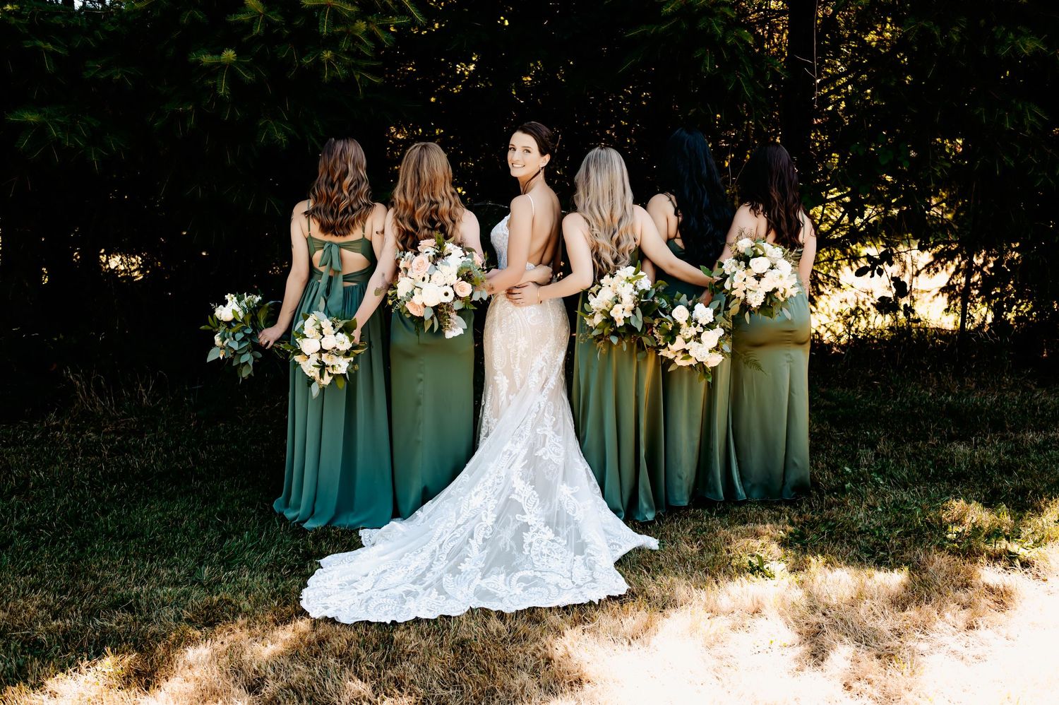 Bride in lace gown stands with bridesmaids in green dresses holding white bouquets outdoors.