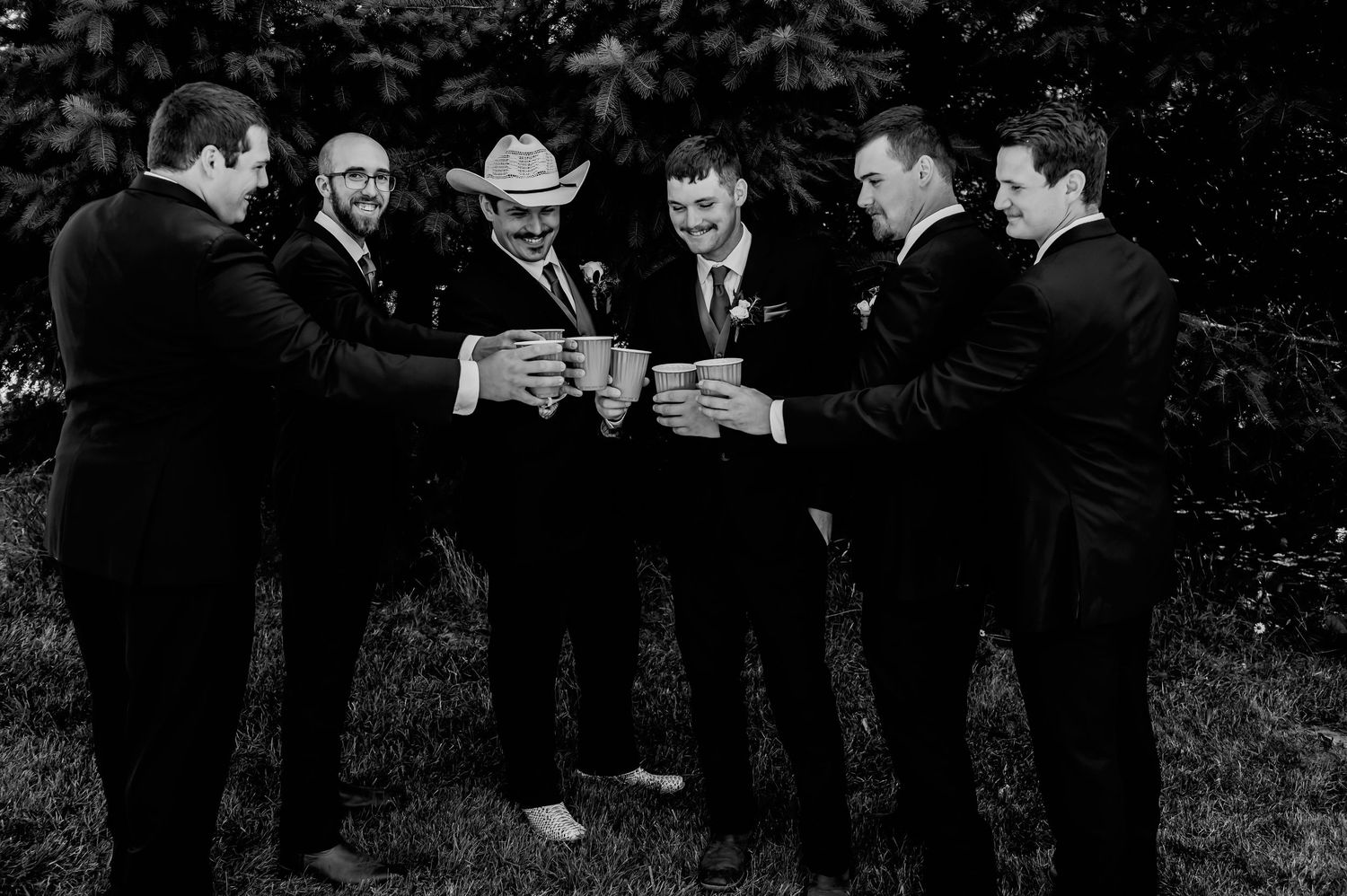 Six groomsmen in dark suits toasting drinks outdoors in a black and white wedding photo.