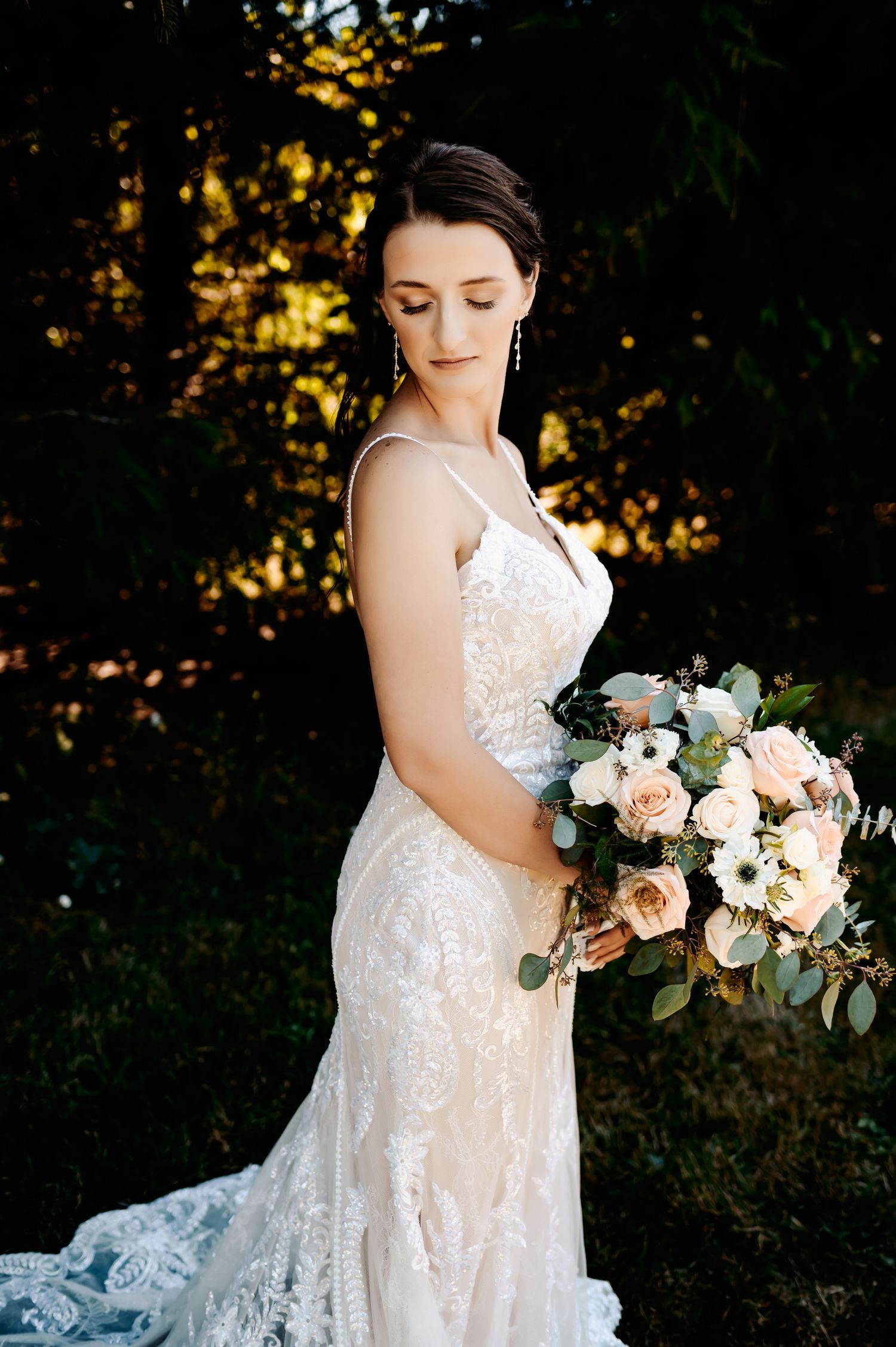Bride in elegant lace wedding gown holding peach and white floral bouquet outdoors.
